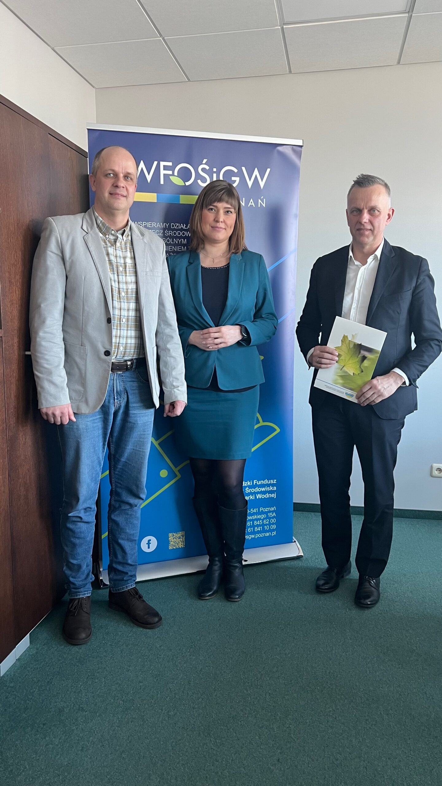 3 osoby stoją na tle ściany i bannera reklamowego.