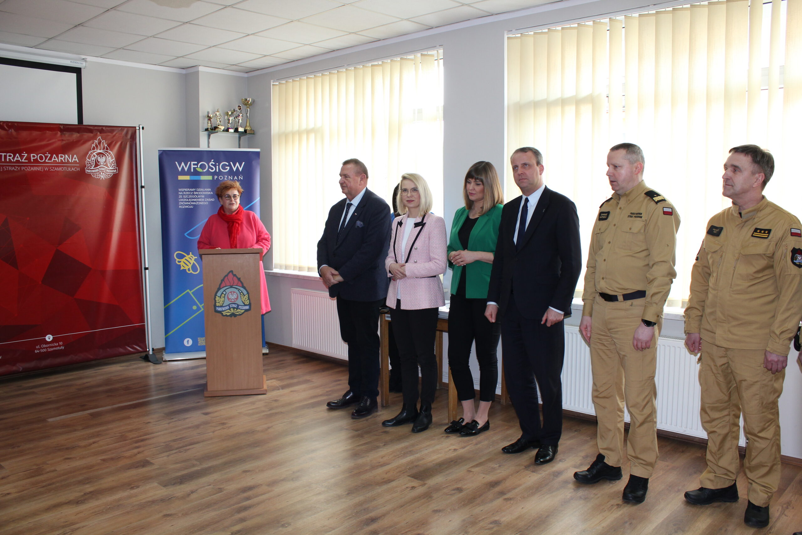 6 osób stoi wzdłuż okien, a 1 osoba przemawia przy mównicy.