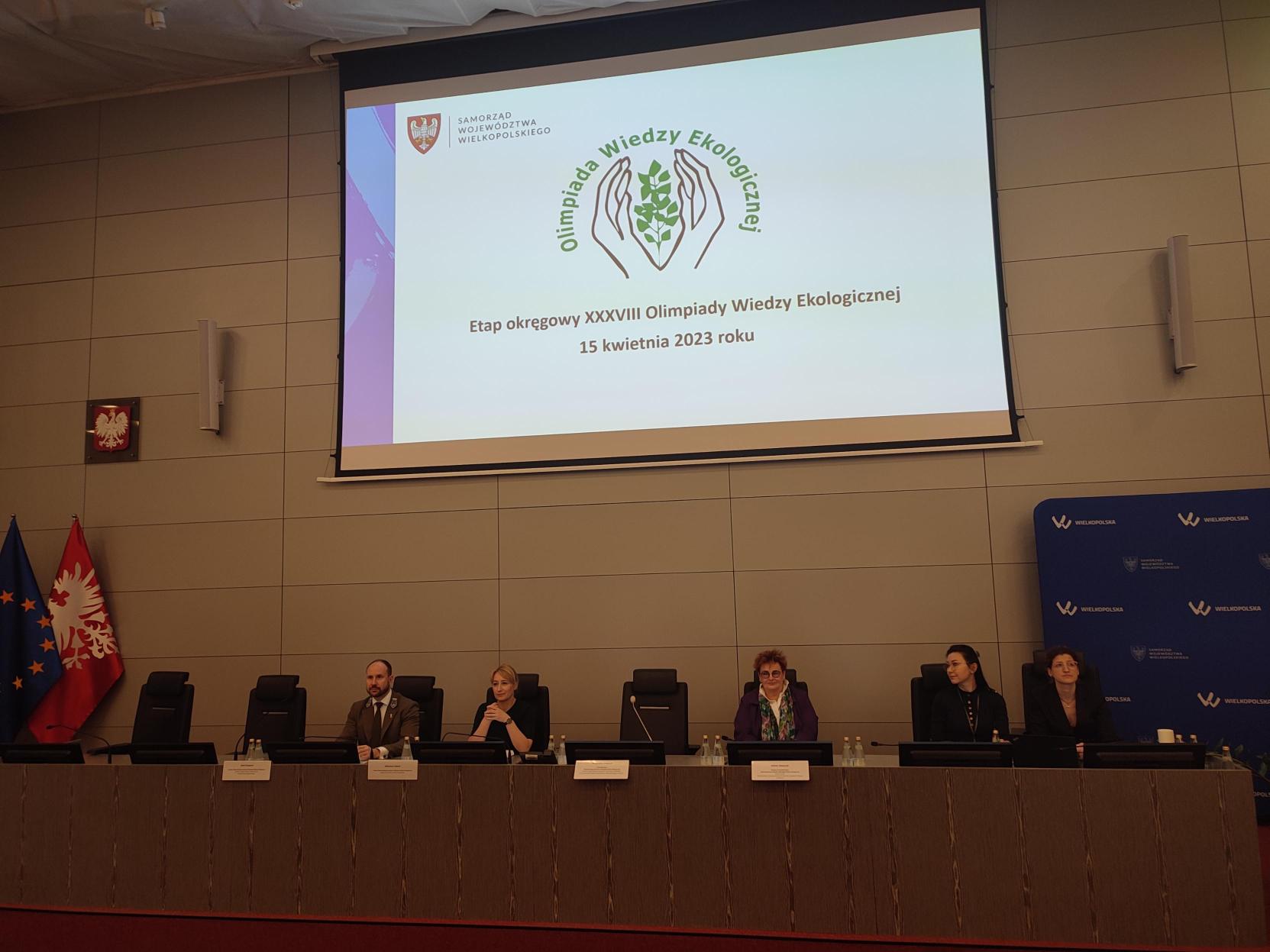 5 osób siedzi przy stole prezydialnym na wielkiej sali.