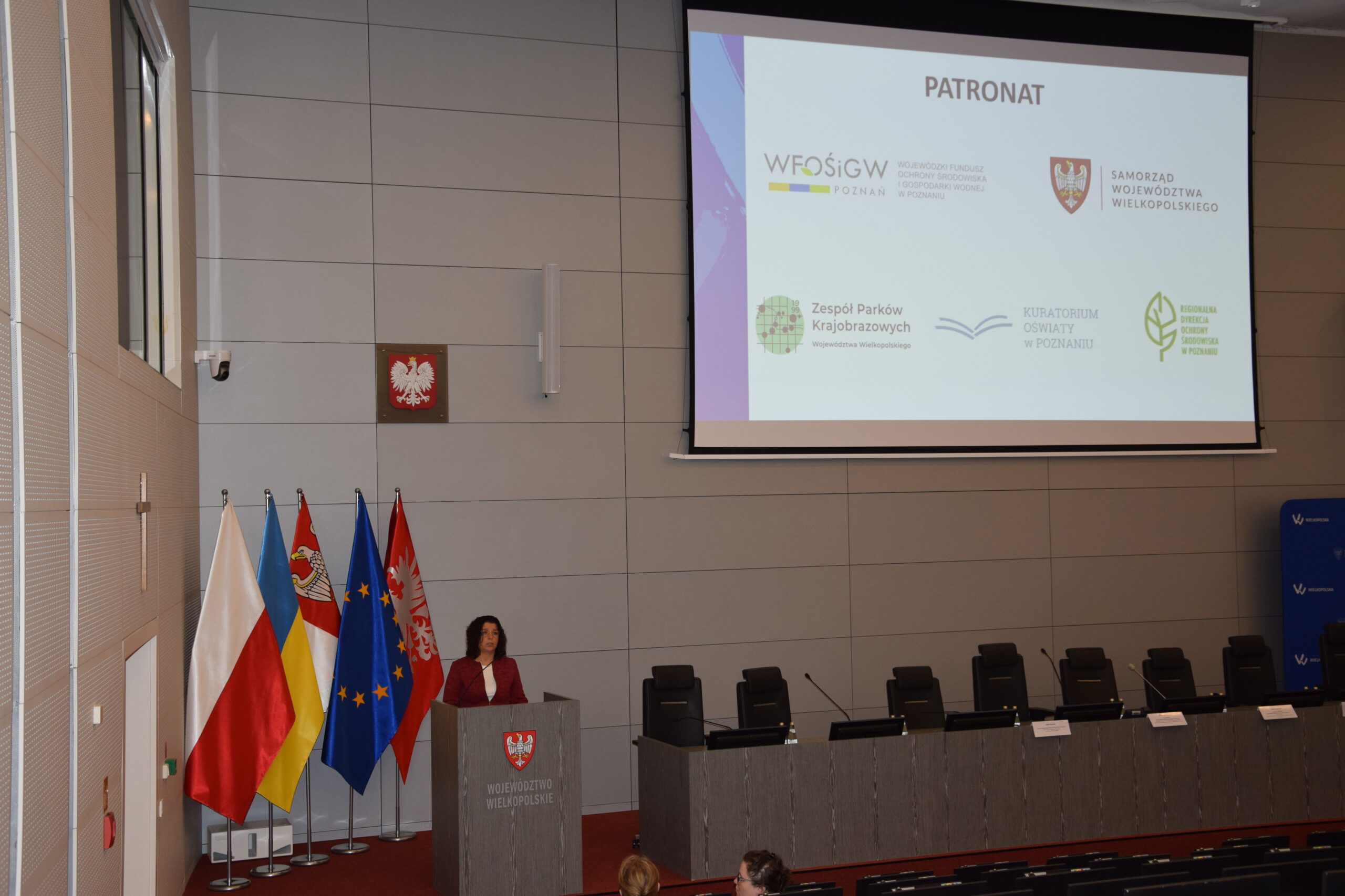 5 osób siedzi przy stole prezydialnym na wielkiej sali, a jedna przemawia przy mównicy.