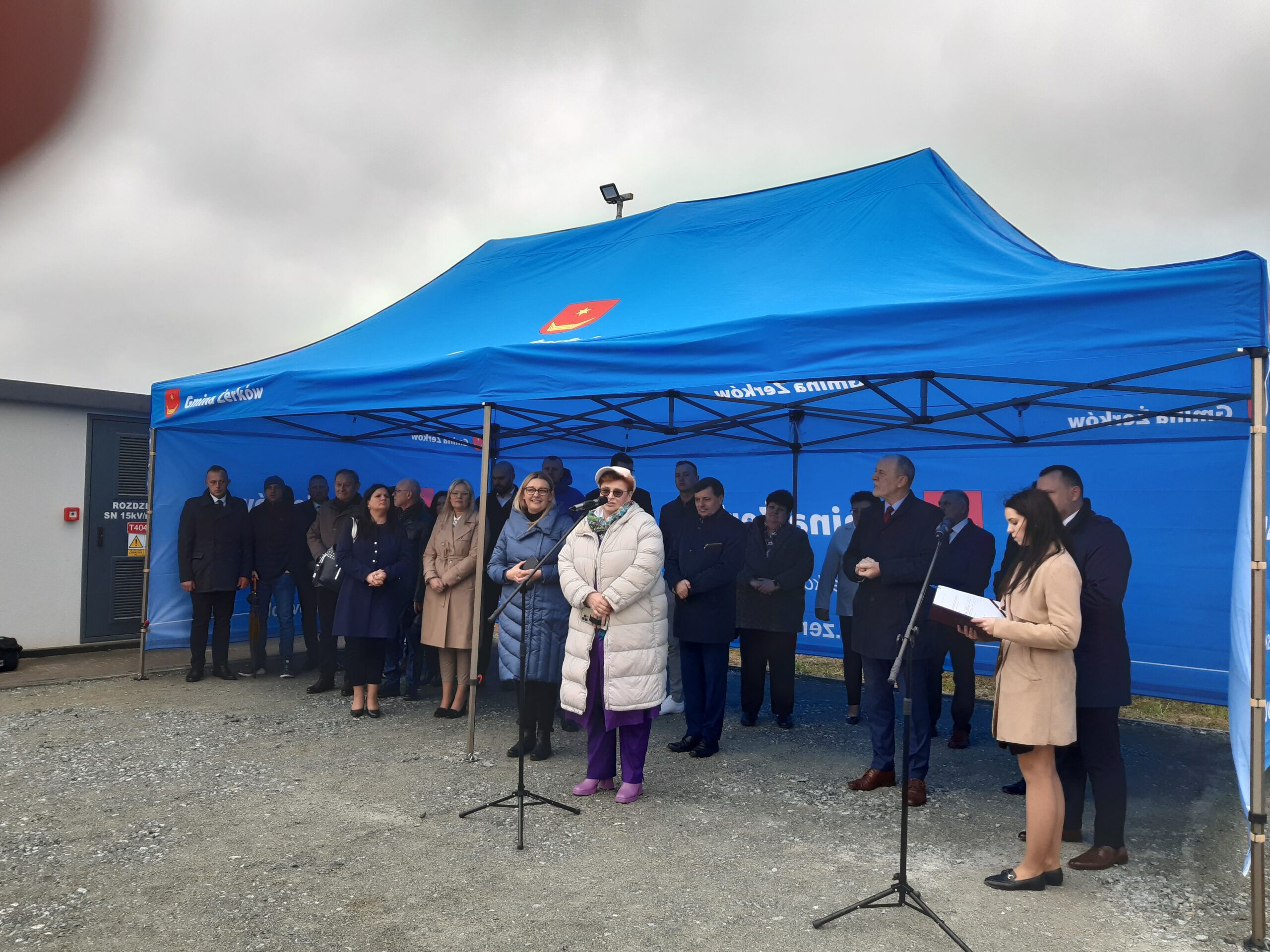 Osoby stoją pod namiotem, a jedna osoba przemawia.