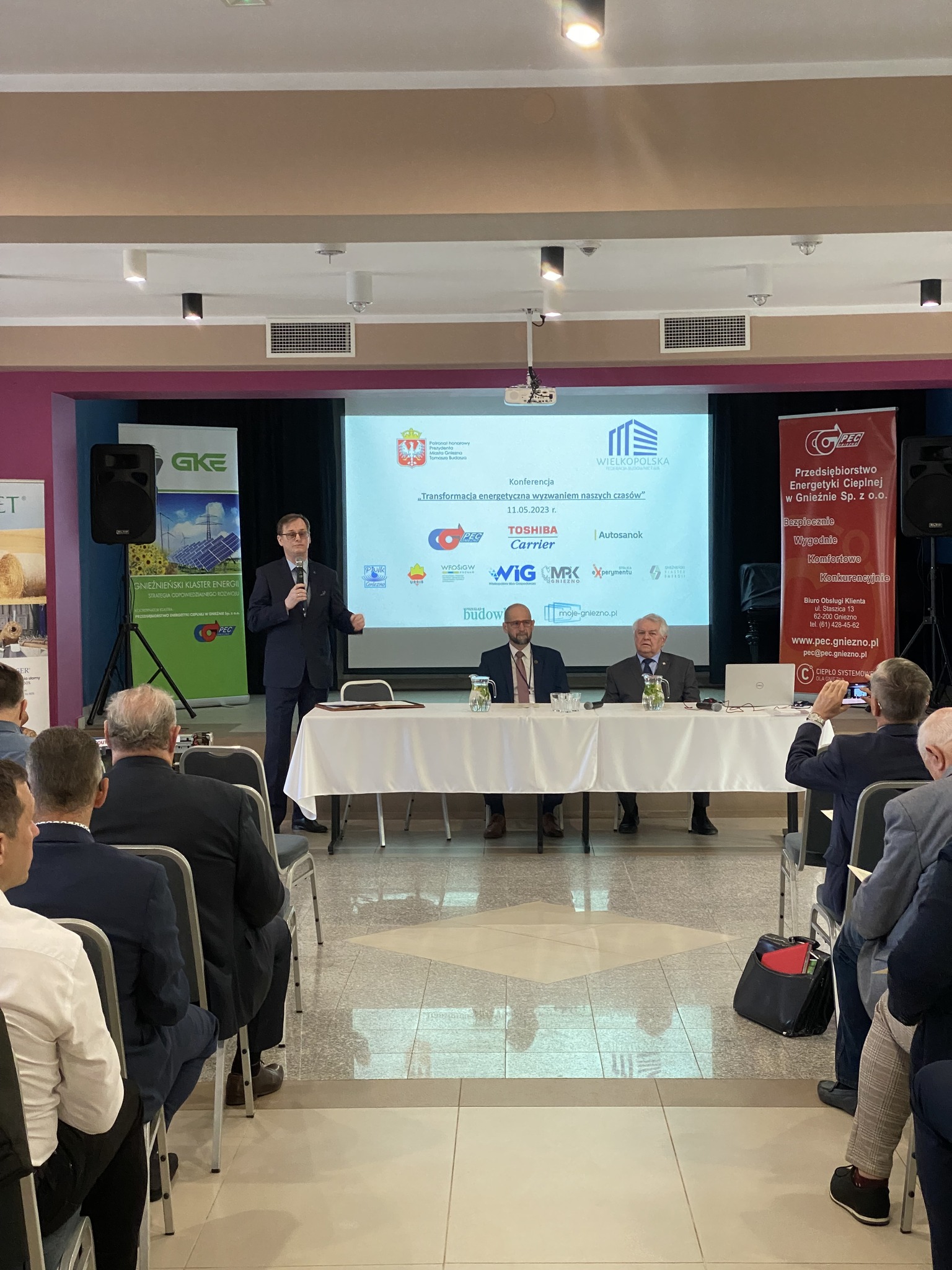 2 osoby siedzą przy stole na tle ściany i prezentacji, a 1 osoba przemawia.