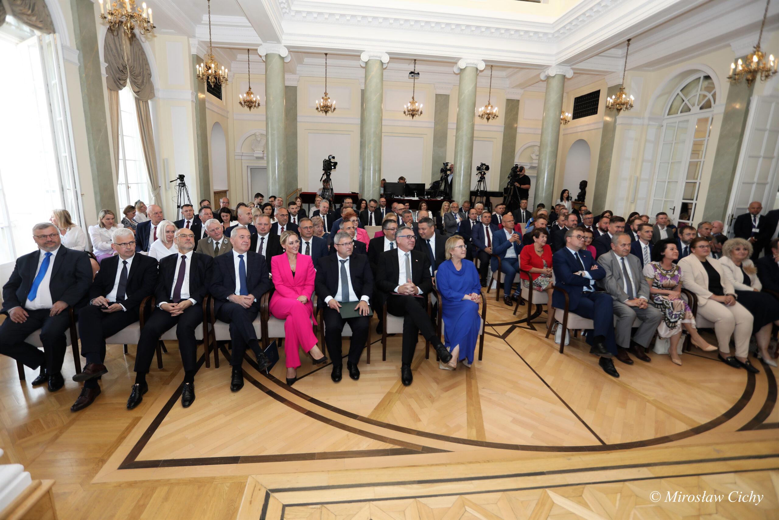 Goście uroczystości siedzą na sali na krzesłach.