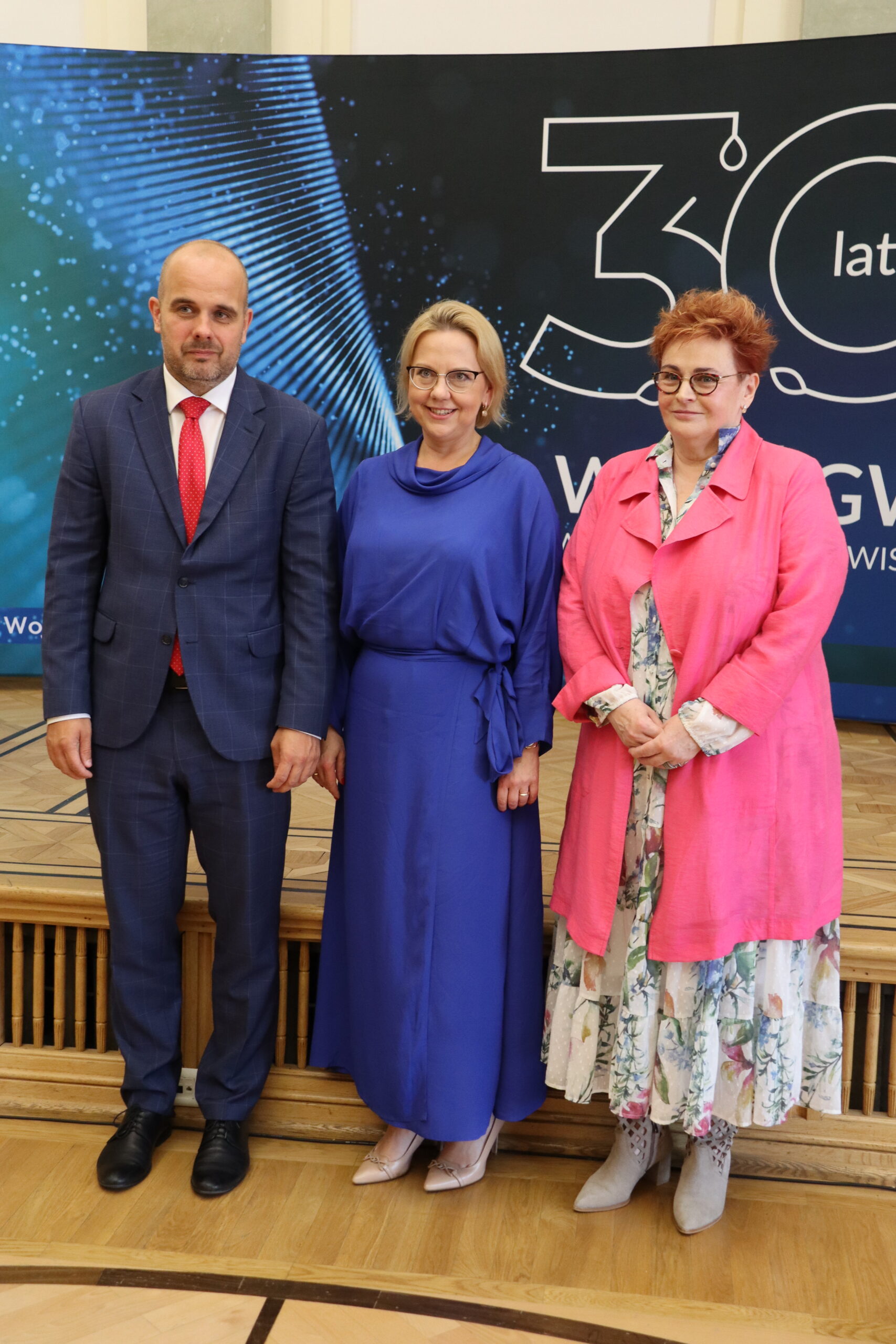 3 osoby stoją na tle ścianki reklamowej