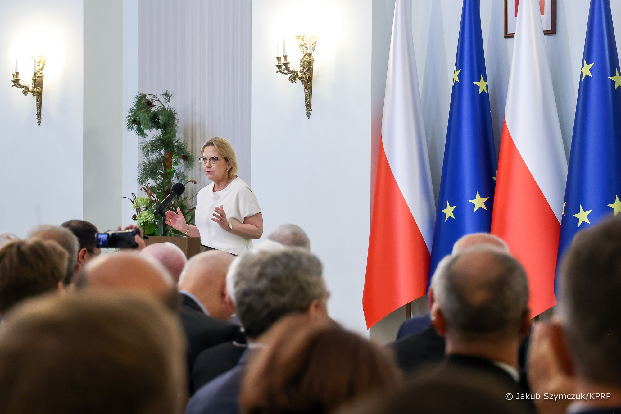  Sala wypełniona ludźmi siedzącymi na krzesłach, a jedna osoba, na środku przemawia stojąc przy mównicy.