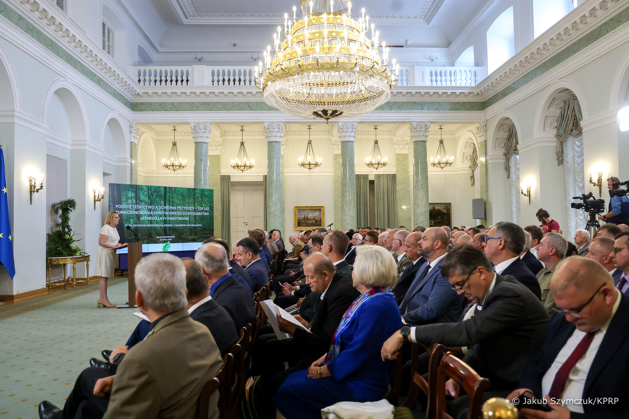  Sala wypełniona ludźmi siedzącymi na krzesłach, a jedna osoba, na środku przemawia stojąc przy mównicy.