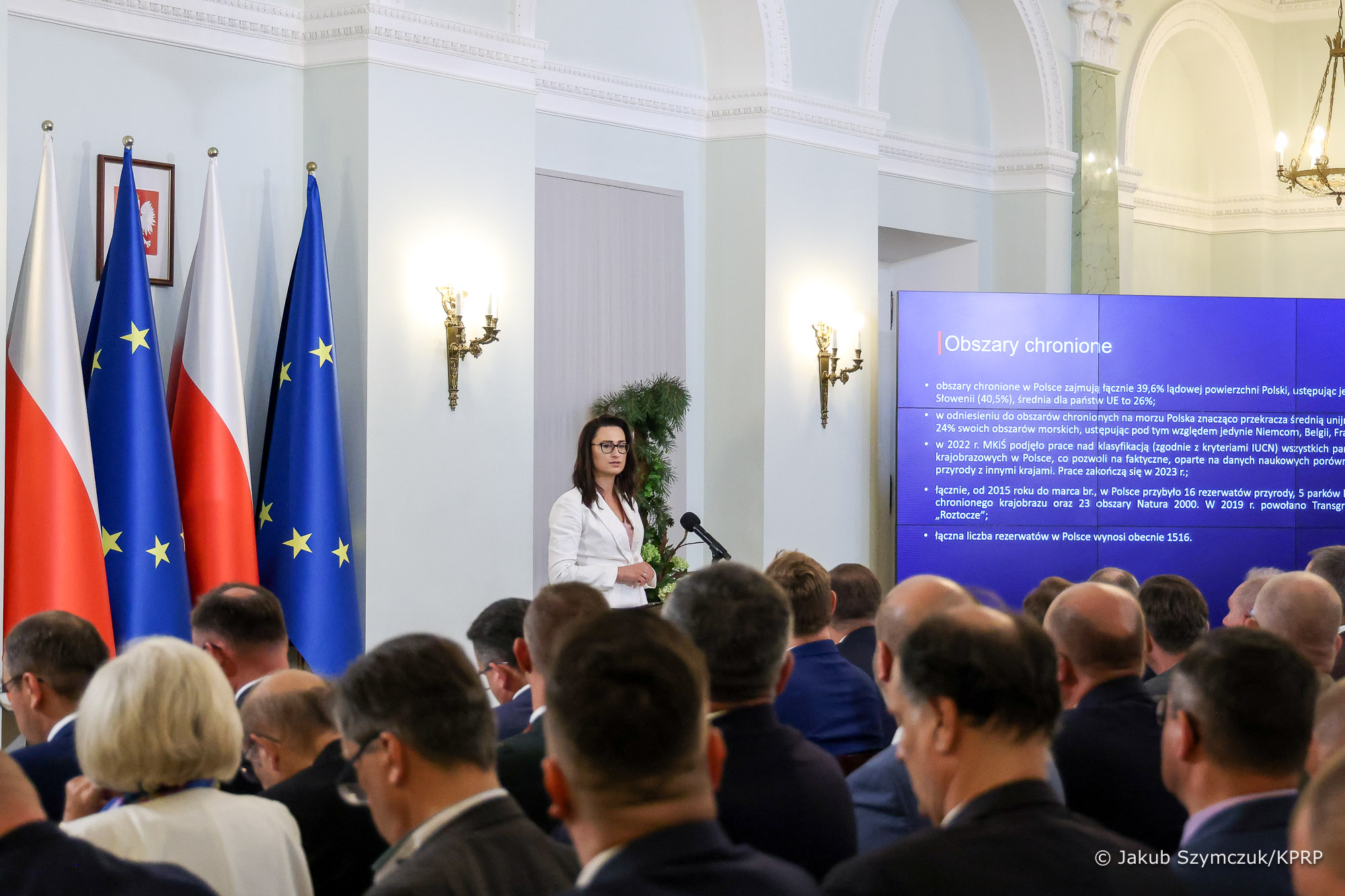  Sala wypełniona ludźmi siedzącymi na krzesłach, a jedna osoba, na środku przemawia stojąc przy mównicy.