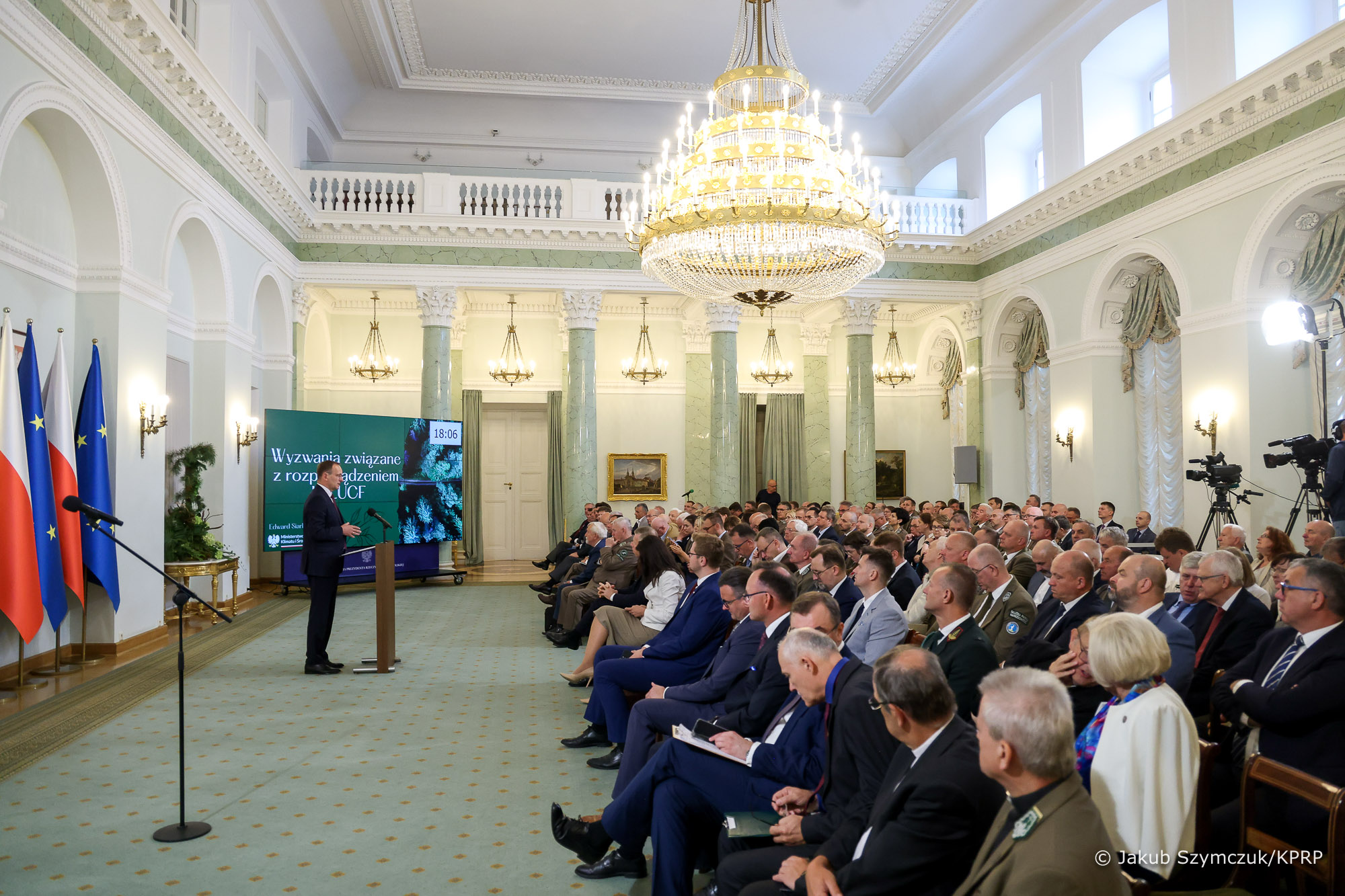  Sala wypełniona ludźmi siedzącymi na krzesłach, a jedna osoba, na środku przemawia stojąc przy mównicy.