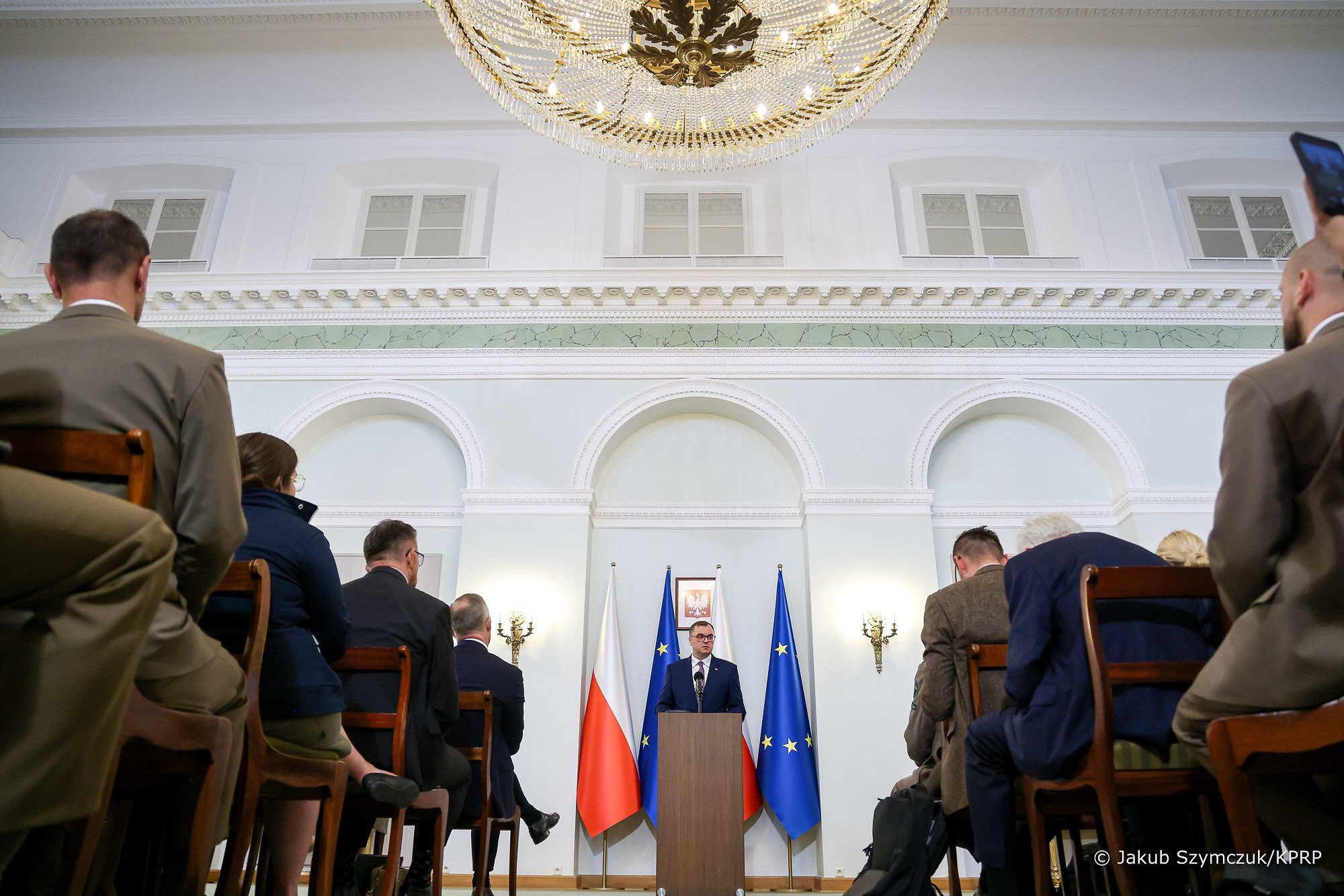  Sala wypełniona ludźmi siedzącymi na krzesłach, a jedna osoba, na środku przemawia stojąc przy mównicy.