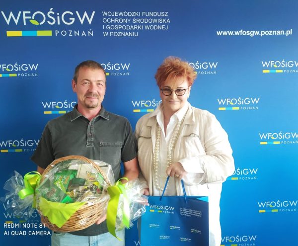 Dwie osoby stoją na tle ścianki reklamowej i pozują, jedna osoba trzyma kosz prezentowy.