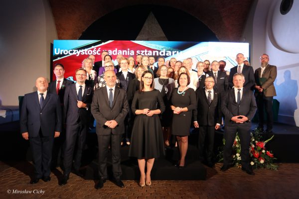 Grupa osób stoi na scenie, na tle telebimu.