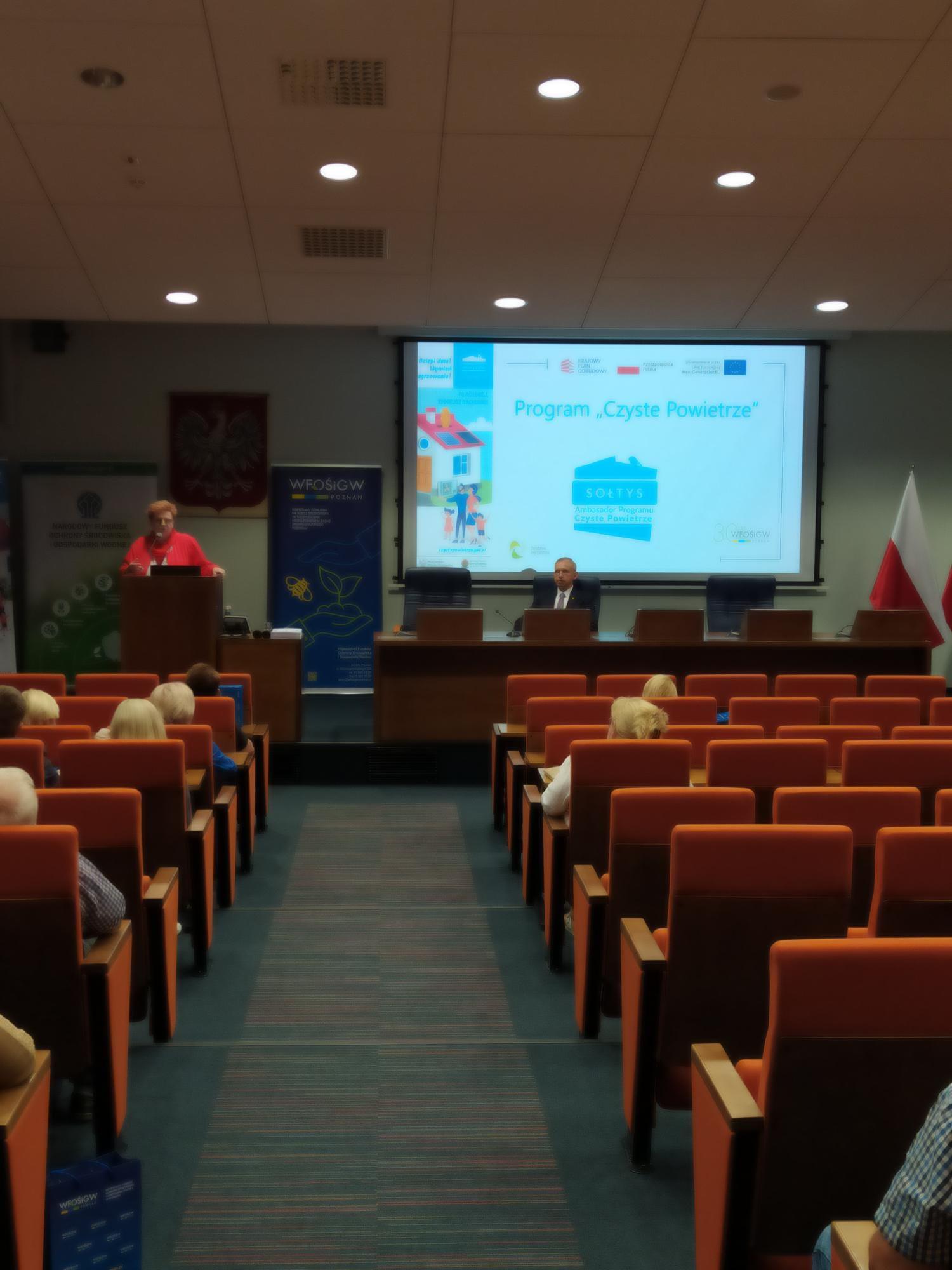 Sala w układzie kinowym, uczestnicy szkolenia siedzą na krzesłach, a 1 osoba przemawia do nich na środku sali,