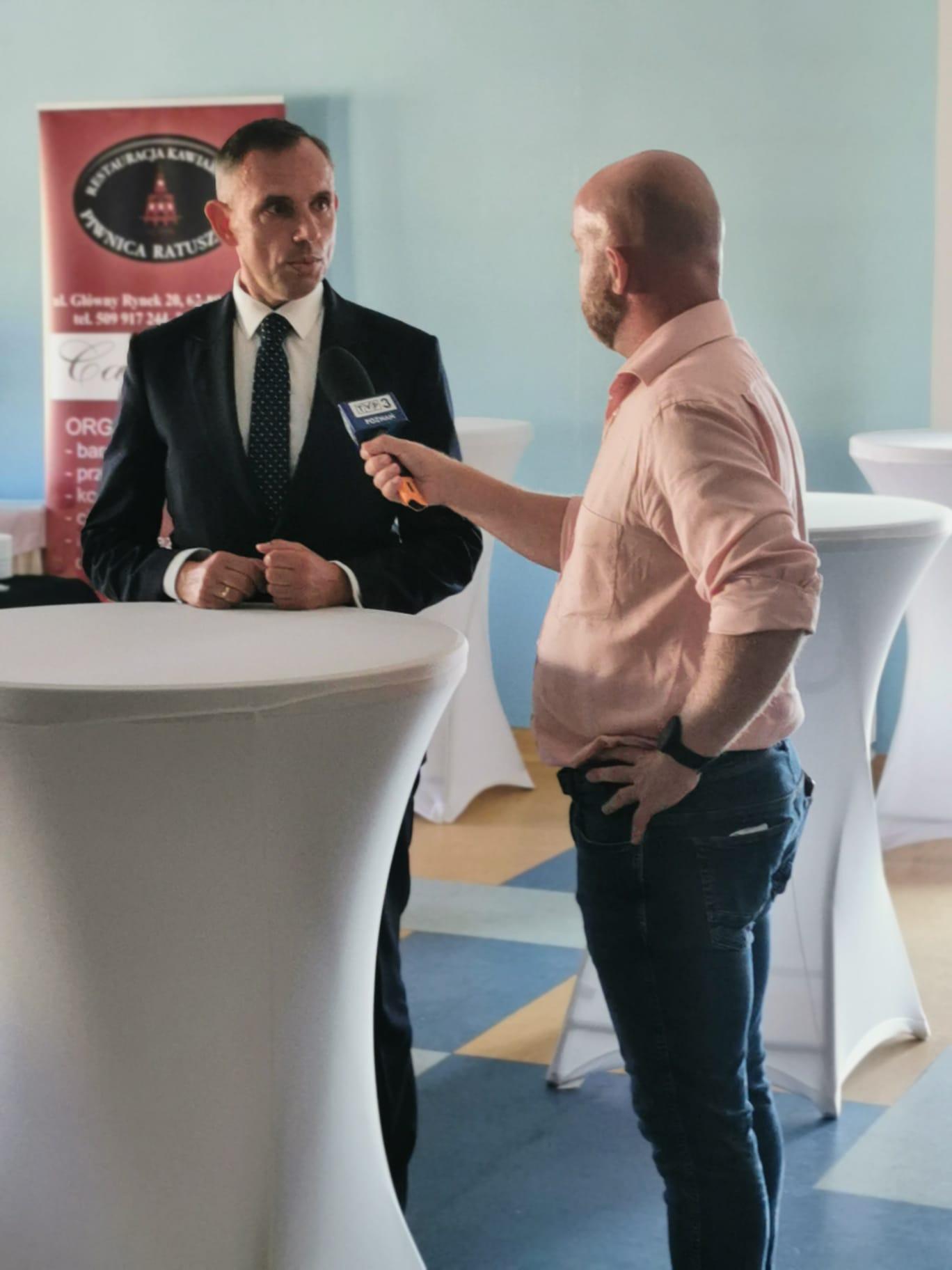 Jedna osoba stoi przystoliku koktajlowym i udziela wywiadu dziennikarzowi.