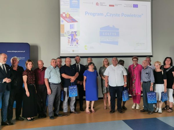 Grupa osób stoi na sali, natle ściany.