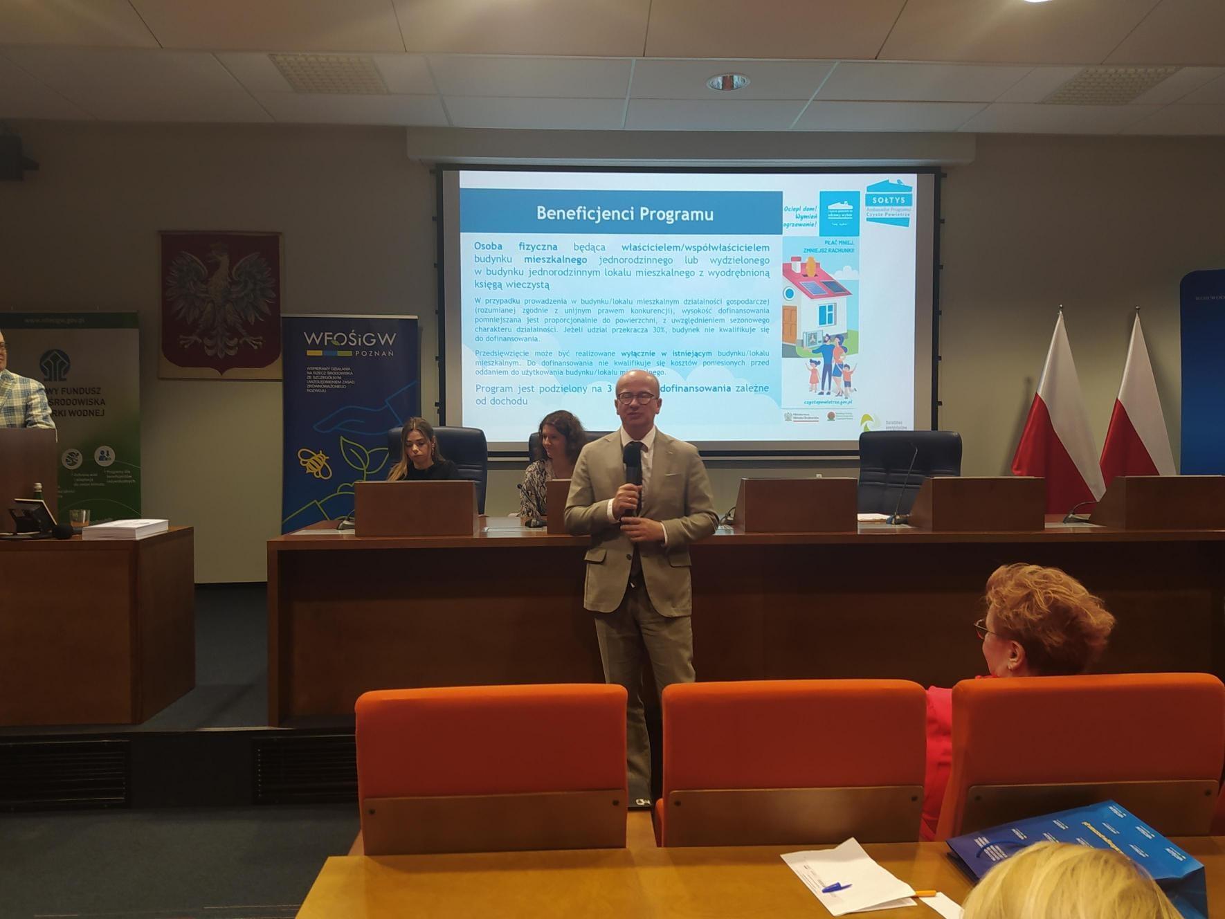 1 osoba stoi na tle ściany z w prezentacją i przemawia.