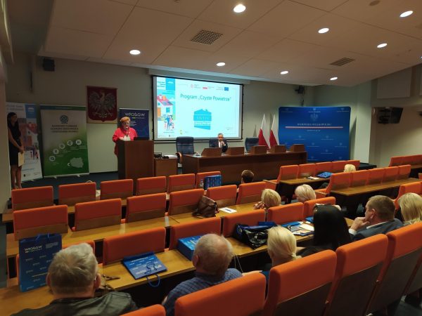 Sala w układzie kinowym, uczestnicy szkolenia siedzą na krzesłach, a 1 osoba przemawia do nich na środku sali,