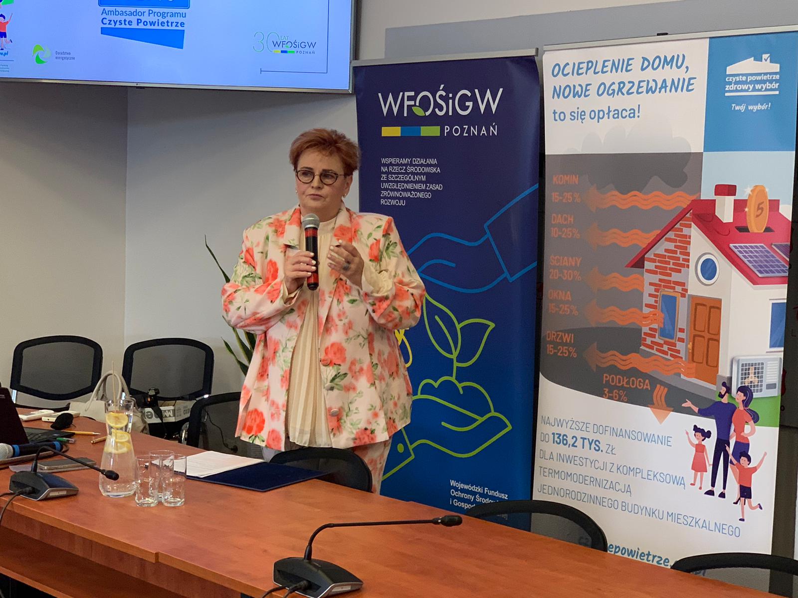 Jedna osoba stoi za stole na tle ściany i przemawia.