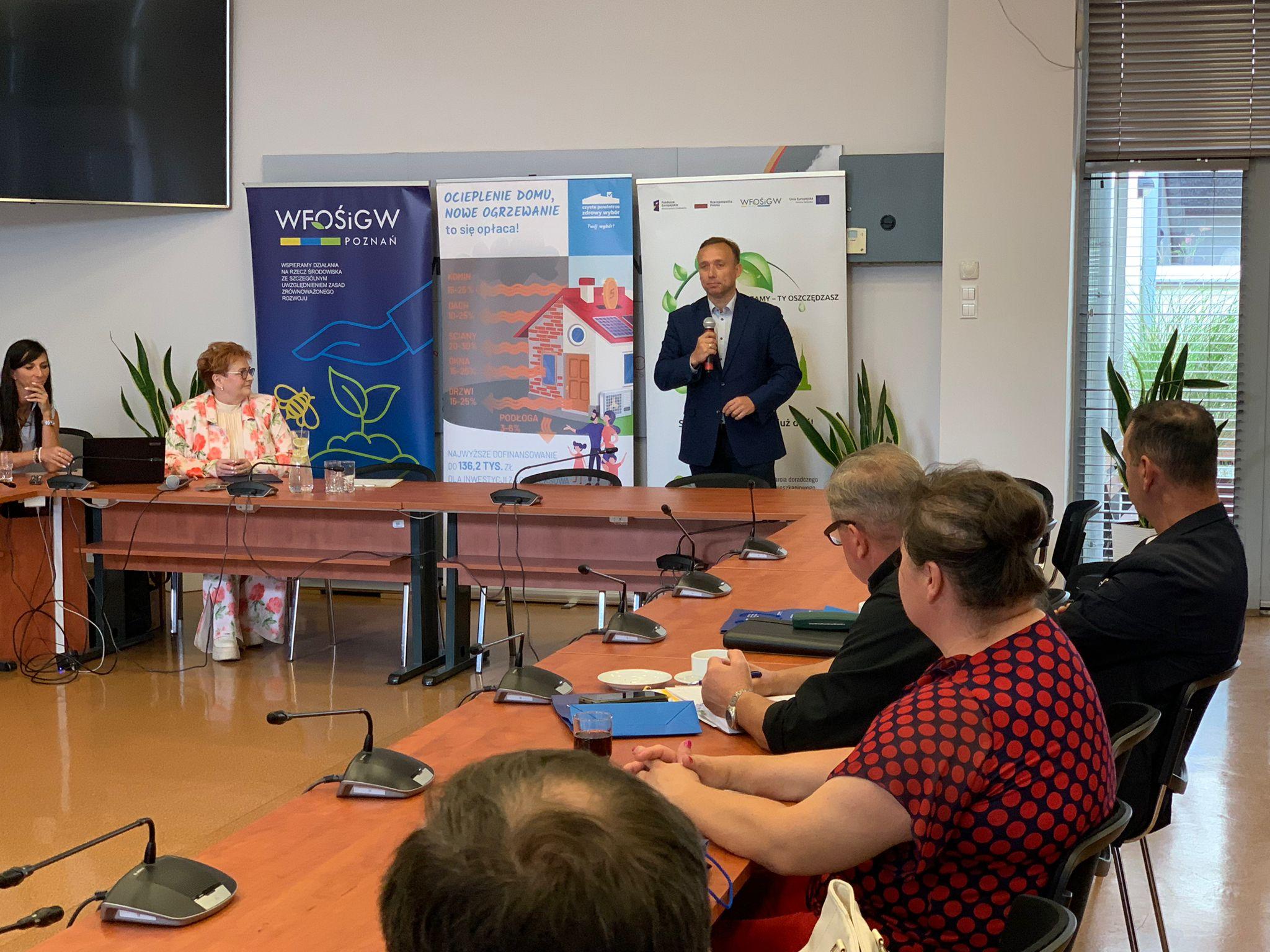 Jedna osoba stoi za stole na tle ściany i przemawia.