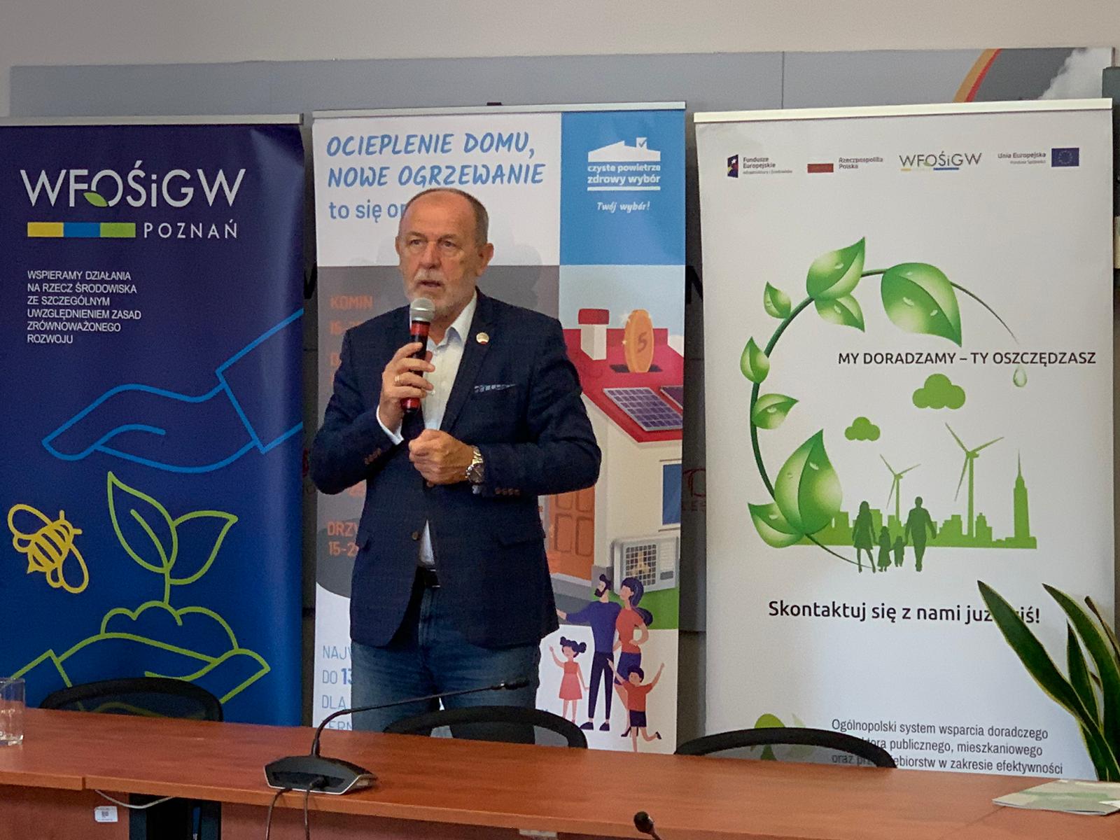 Jedna osoba stoi za stole na tle ściany i przemawia.