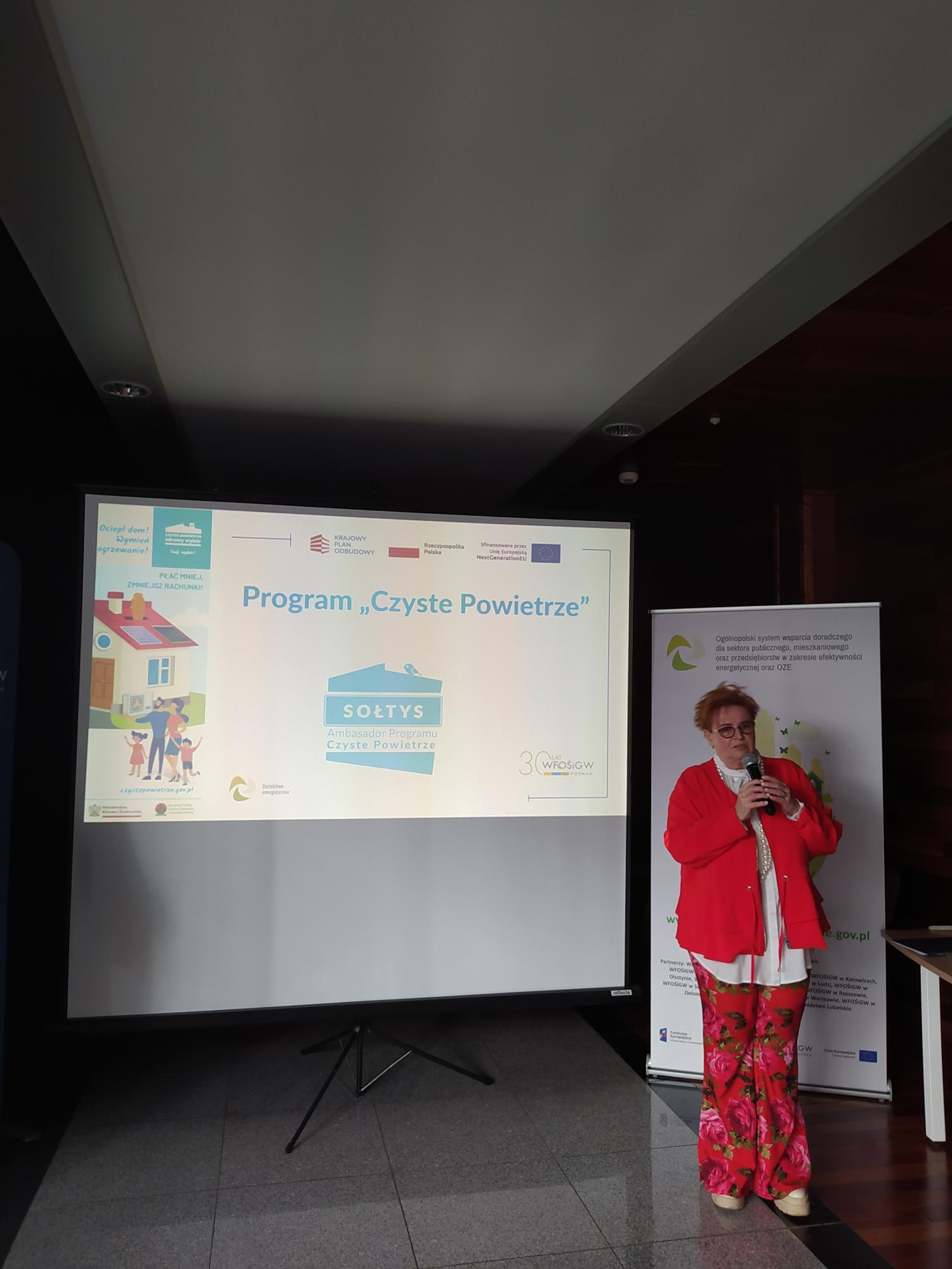 Jedna osoba stoi na tle prezentacji i przemawia do osób siedzących na sali.