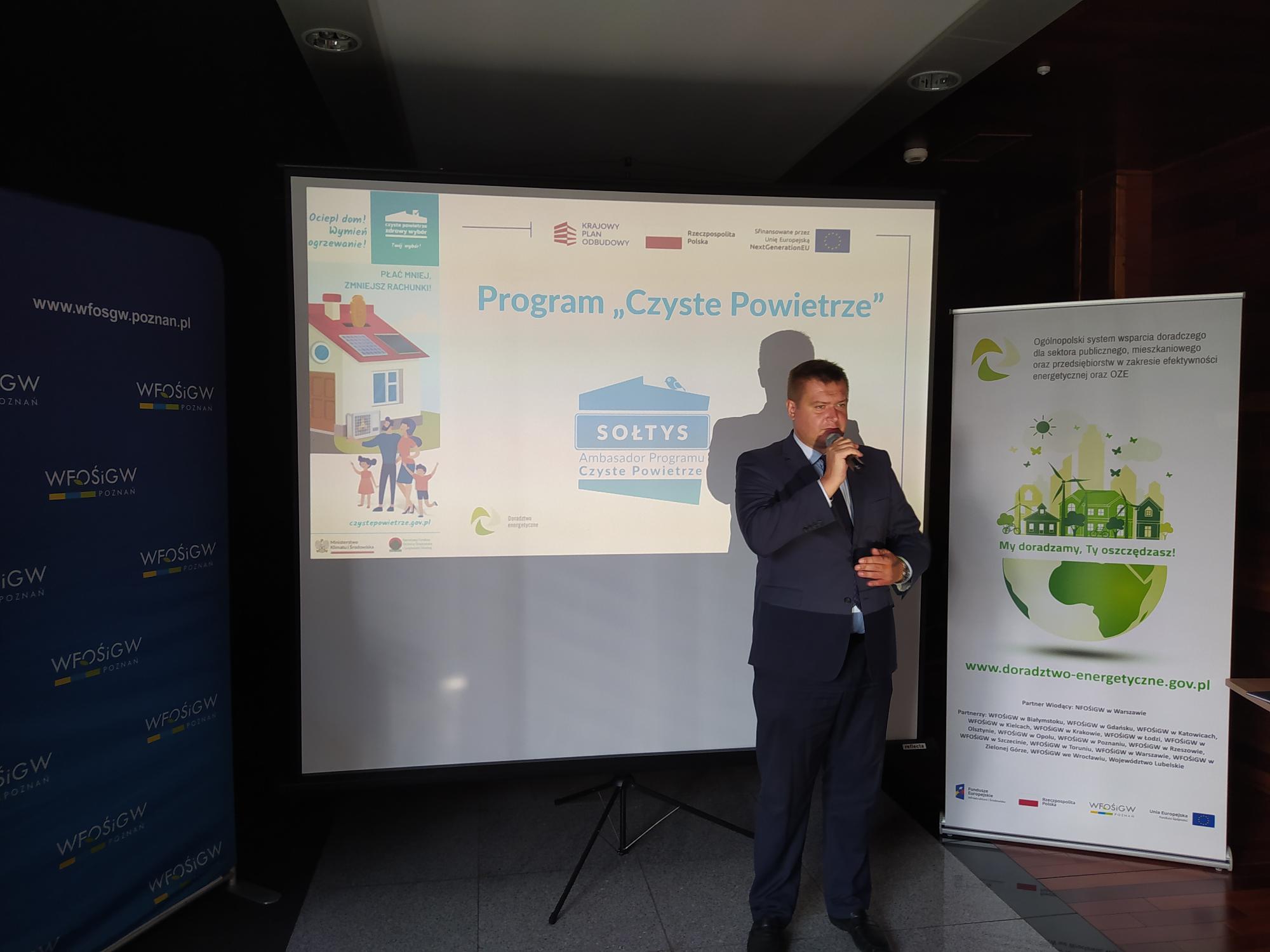 Jedna osoba stoi na tle prezentacji i przemawia do osób siedzących na sali.