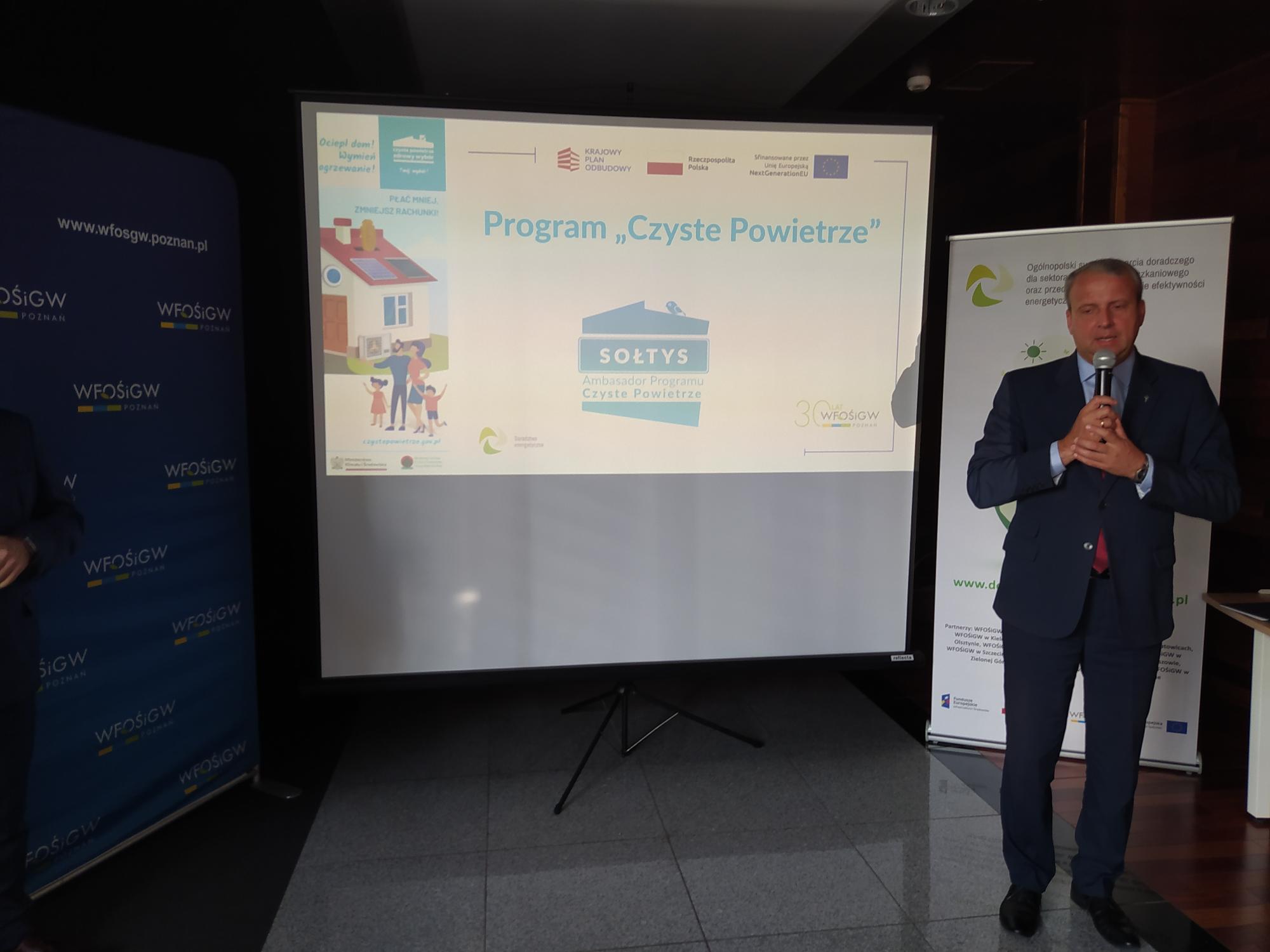 Jedna osoba stoi na tle prezentacji i przemawia do osób siedzących na sali.