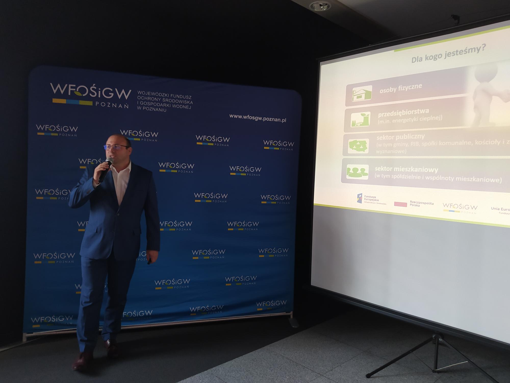 Jedna osoba stoi na tle prezentacji i przemawia do osób siedzących na sali.