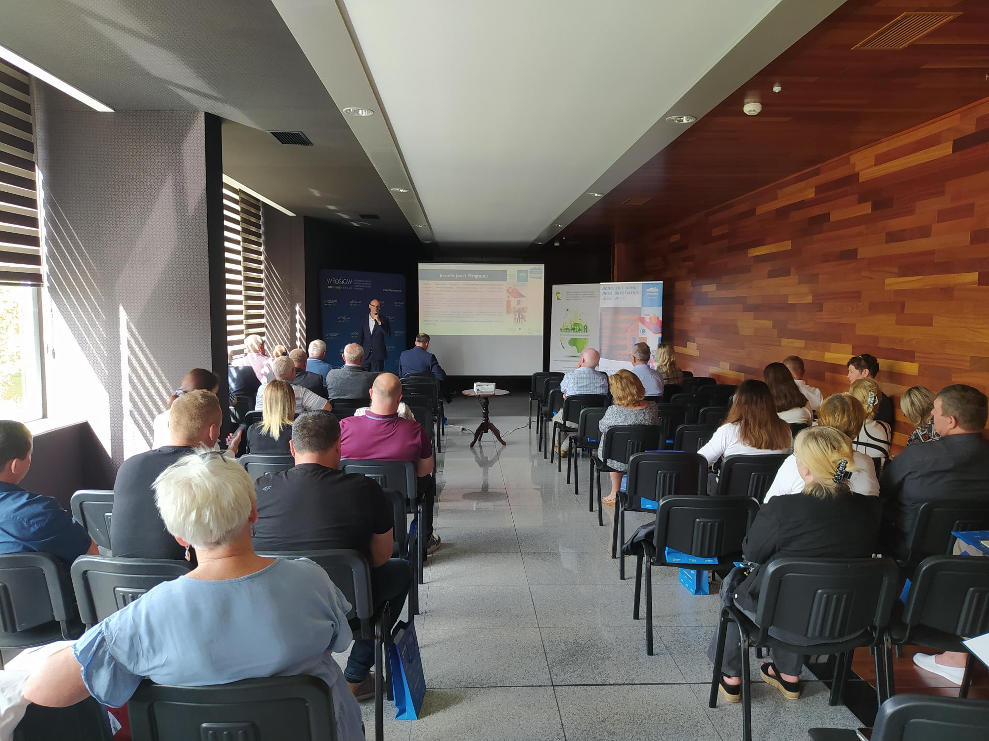 Ludzie siedzą na krzesłach w sali i słuchają prezentacji.