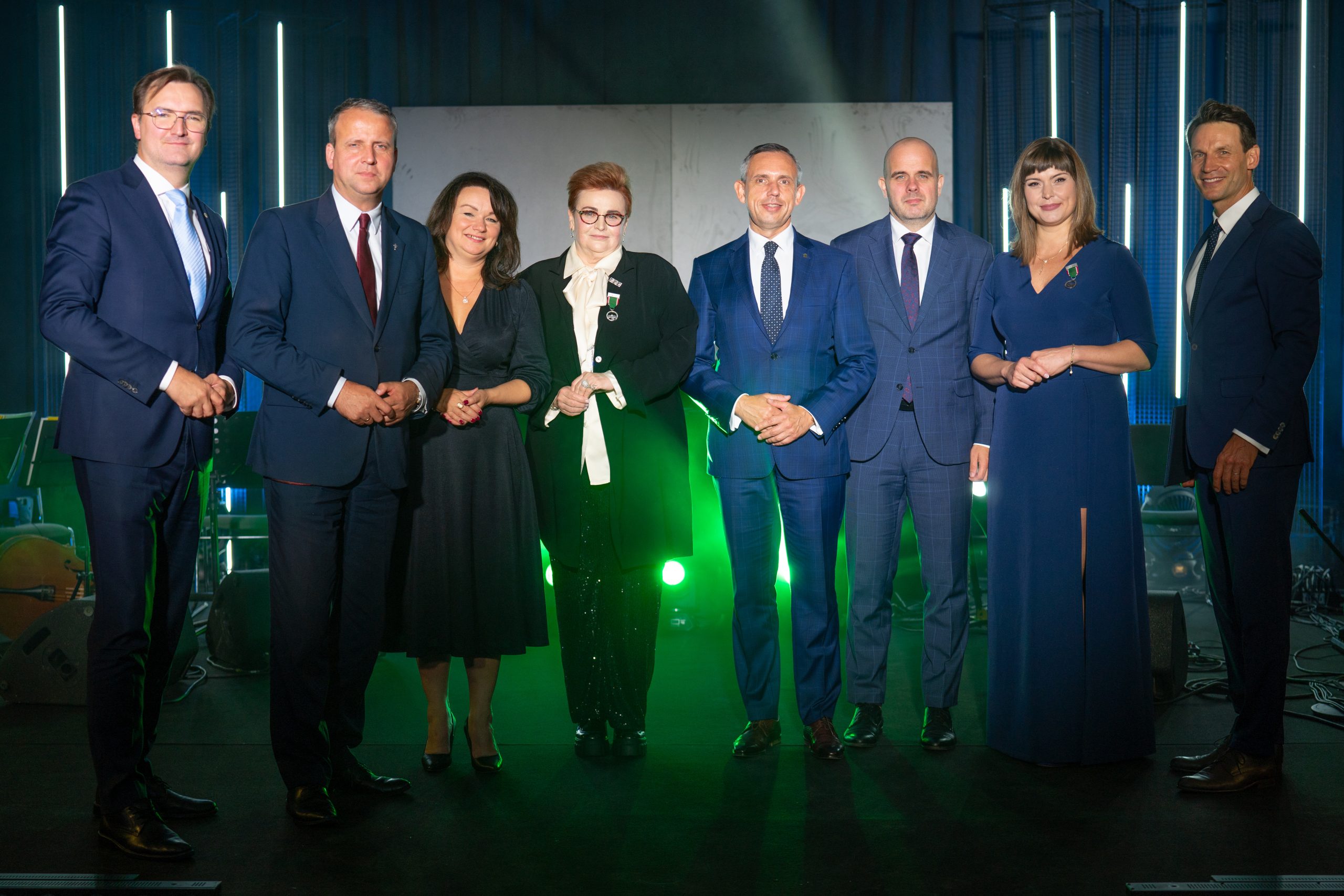 8 osób stoi na scenie w blasku światła i pozuje do wspólnego zdjęcia.