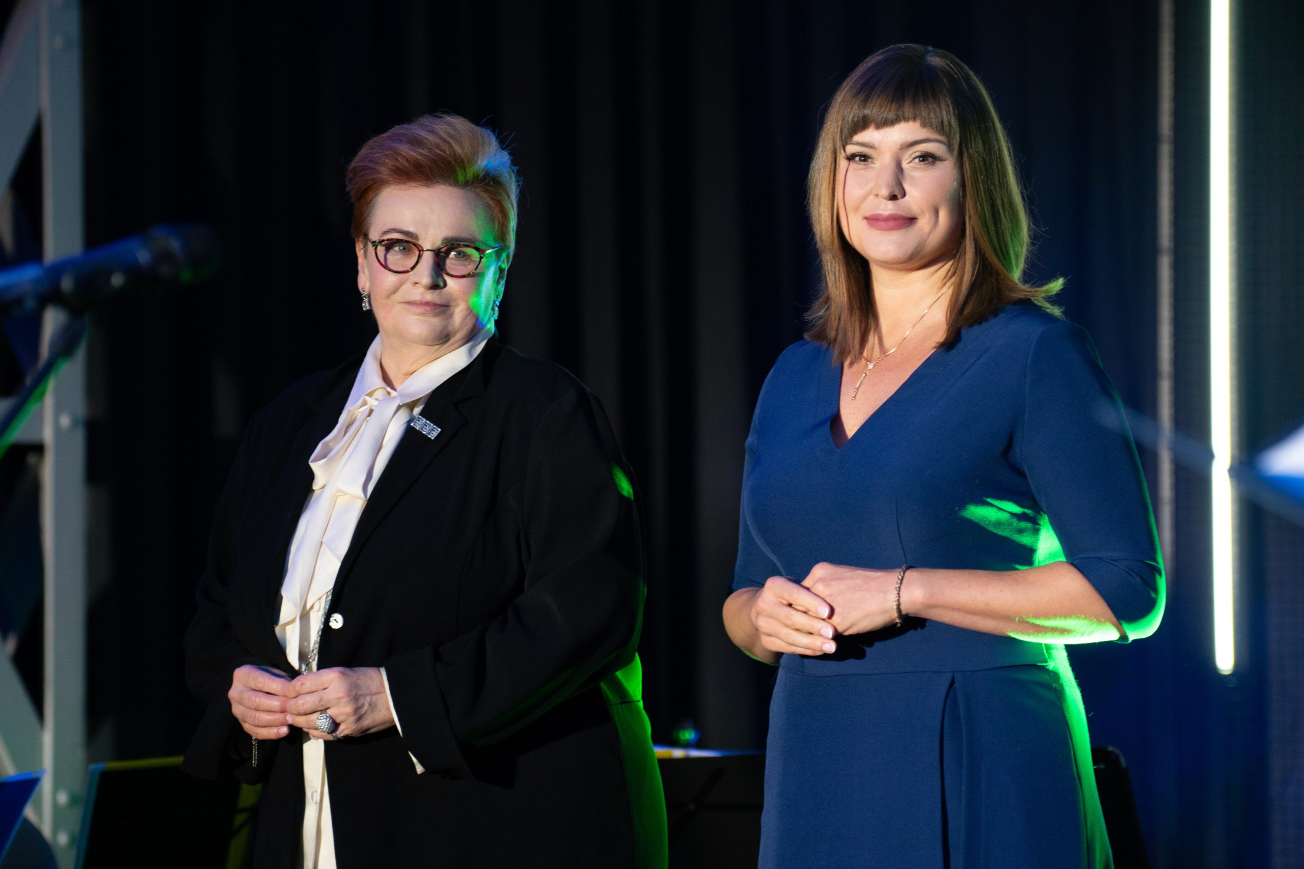 2 osoby stoją na scenie i spoglądają w kierunku obiektywu.