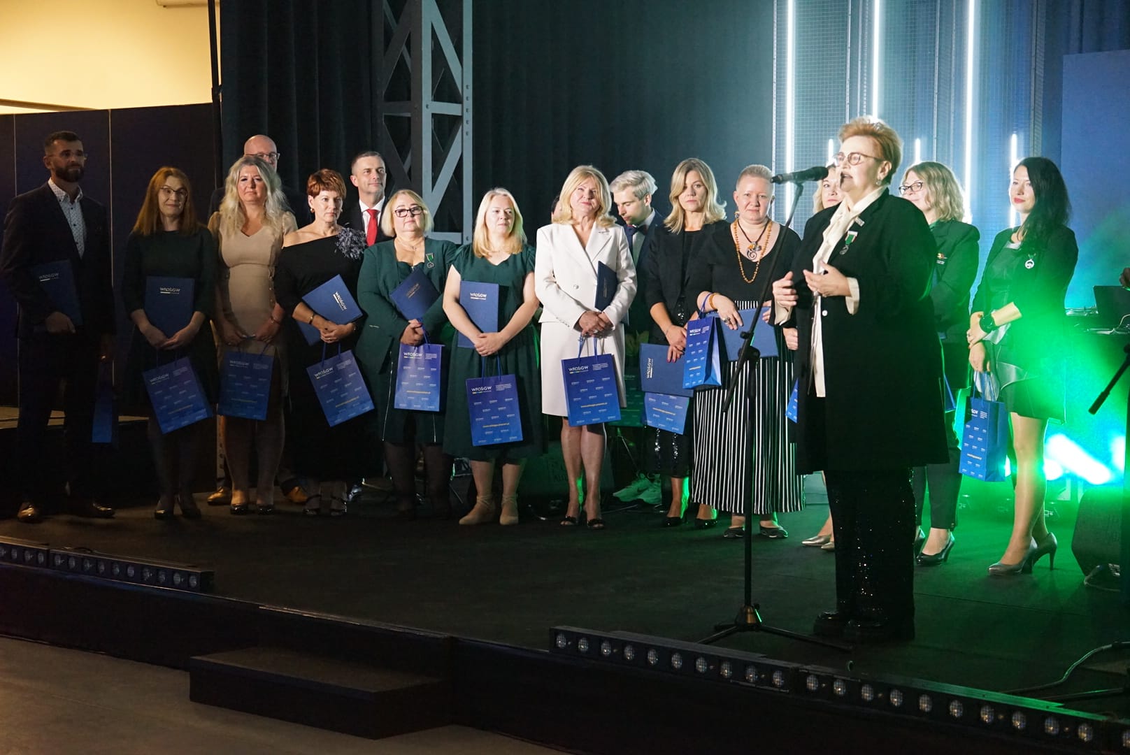 Duża grupa osób stoi na scenie w blasku światła i pozuje do wspólnego zdjęcia.