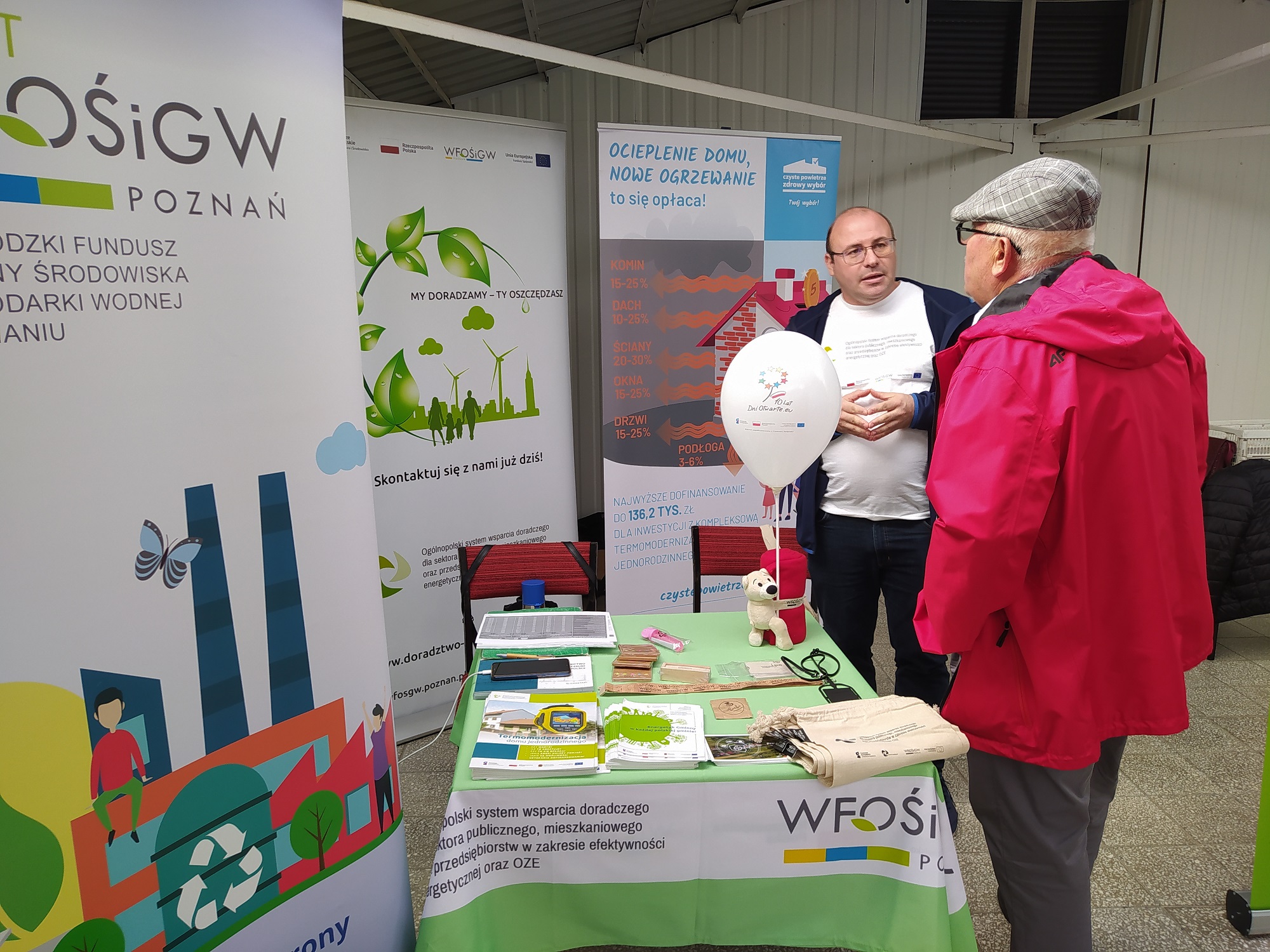 3 osoby stoją na tle bannerów reklamowych i stoiska.