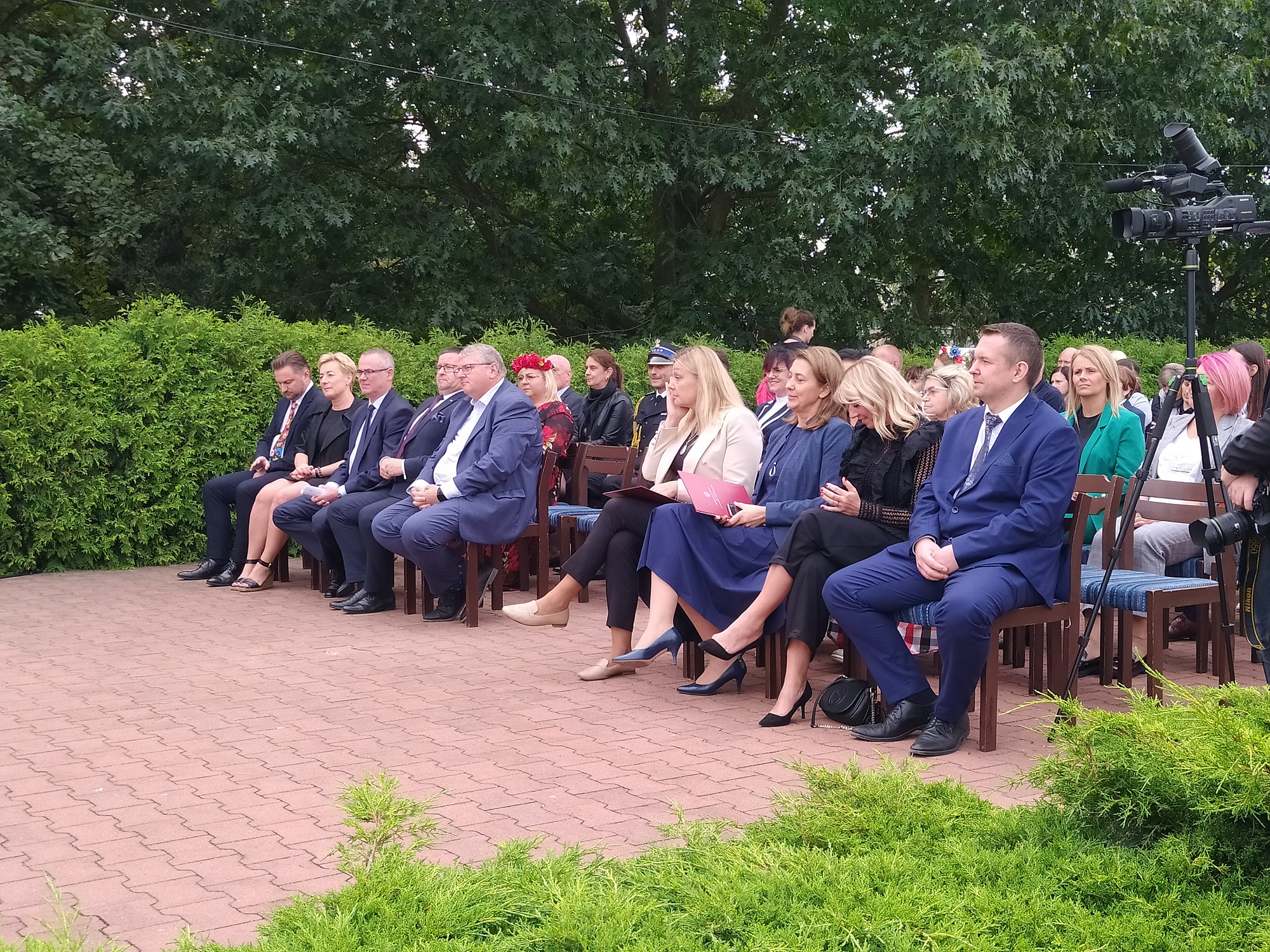 Grupa osób siedzi na krzesłach przed sceną.