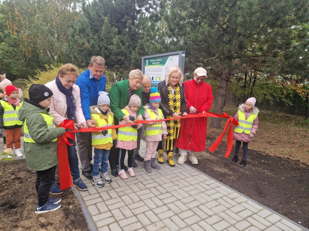 Grupa osób stojaca na początku ścieżki edukacyjnej, dorosli oraz dzieci - tuż przed czerwoną wstęgą, którą będą uroczyście przecinać.