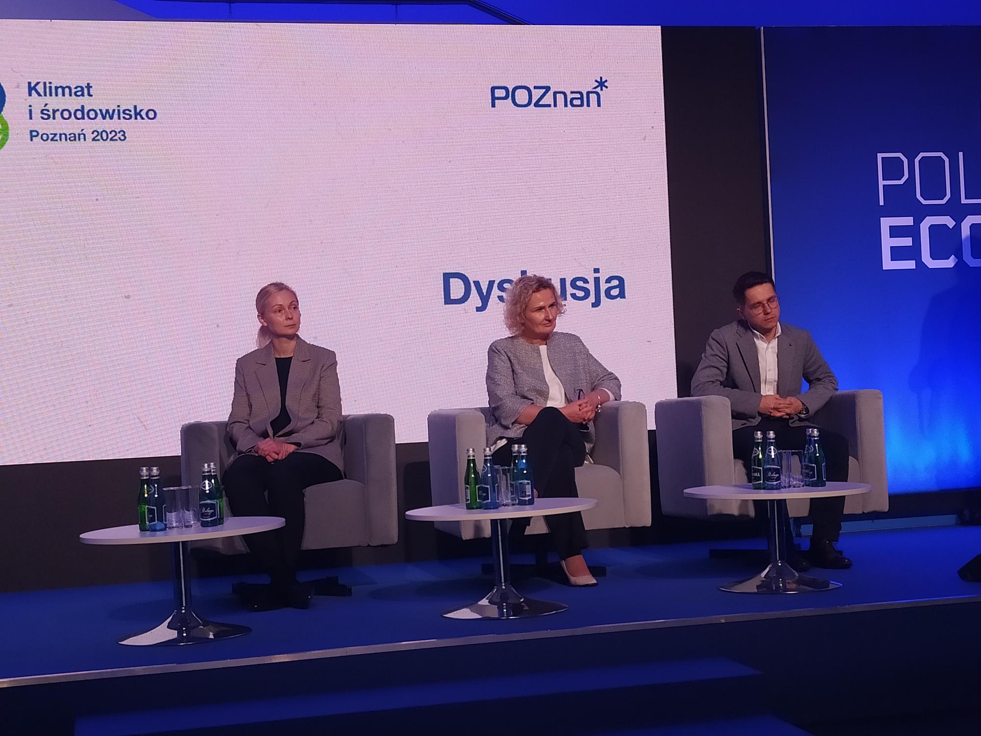 3 osoby siedzą na scenie i debatują podczas konferencji.