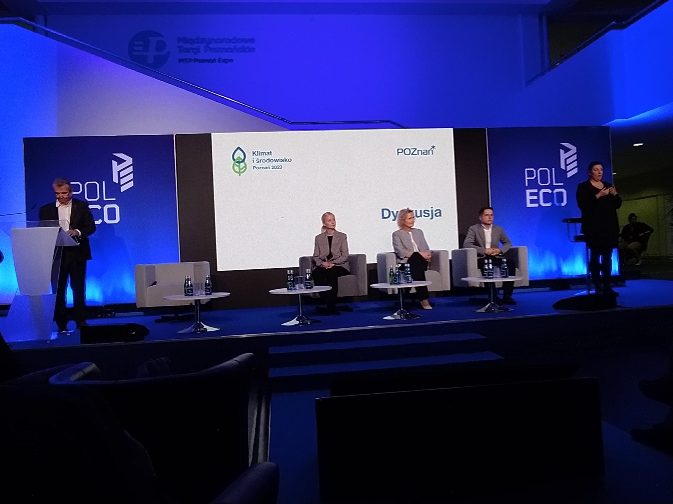 3 osoby siedzą na scenie i debatują podczas konferencji.