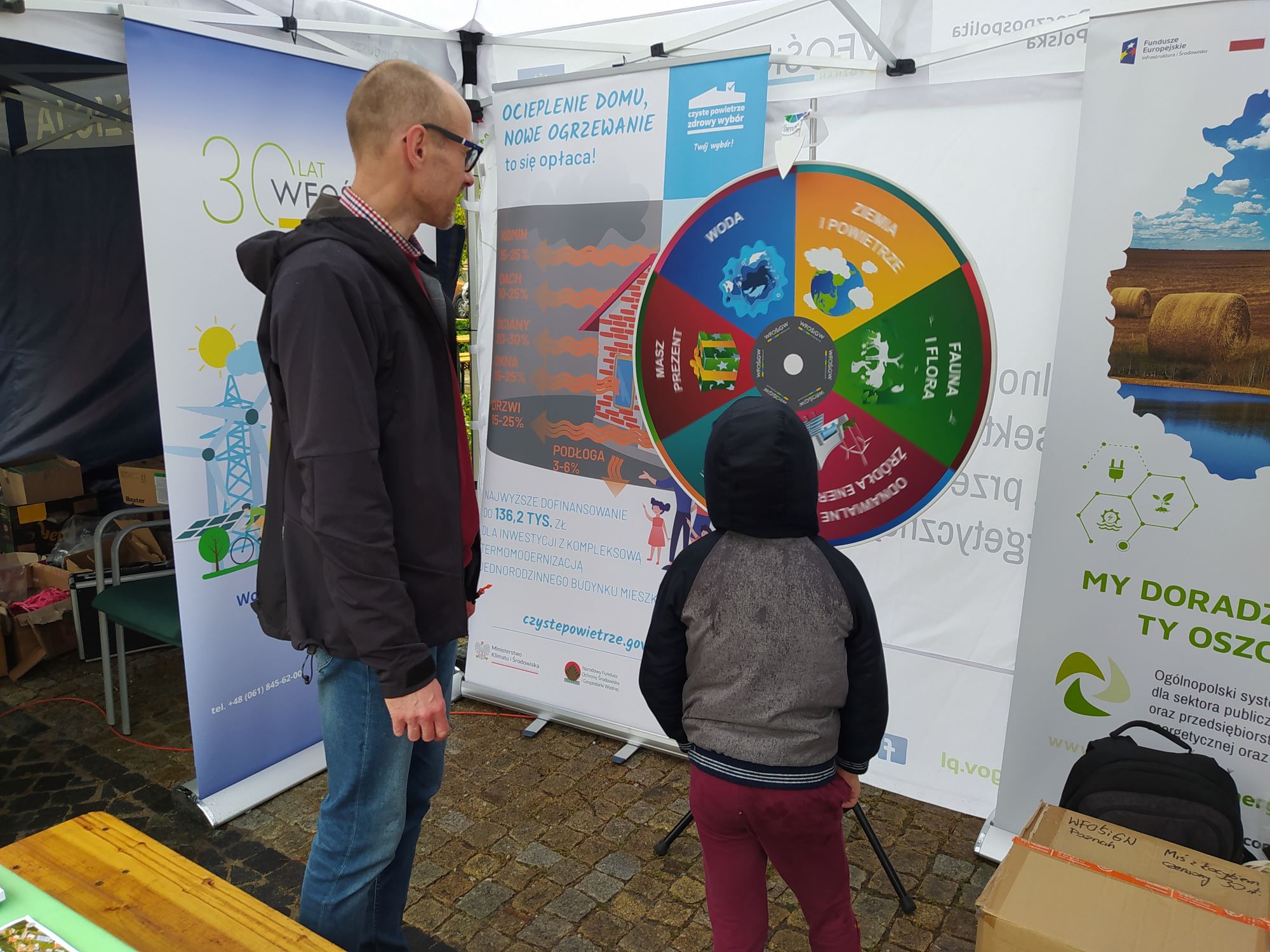 Pod namiotem stoją 2 osoby, mały chłopiec gra w ekologiczne koło fortuny