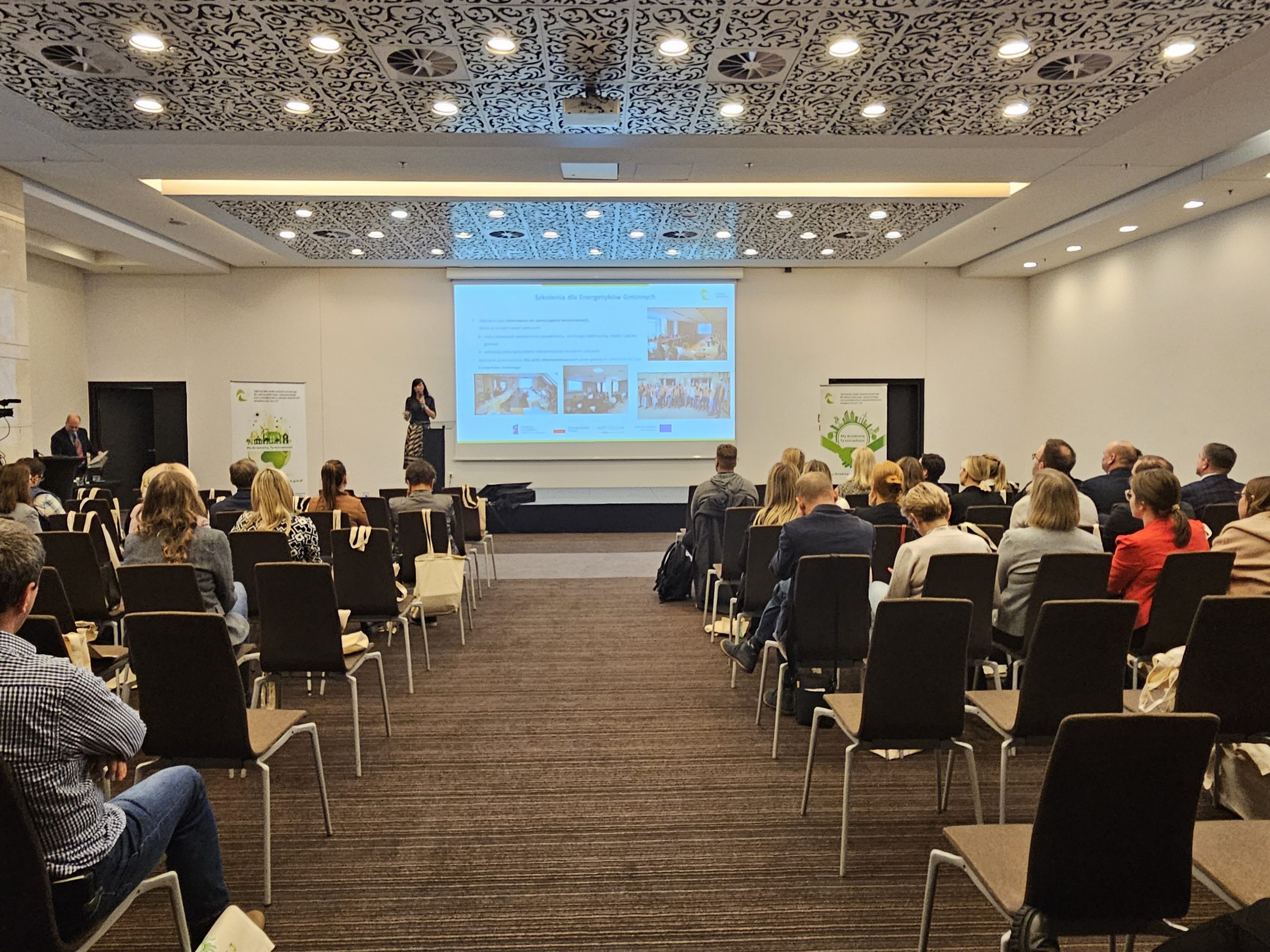 1 osoba stoi na scenie w trakcie konferencji i przemawia do uczestników.