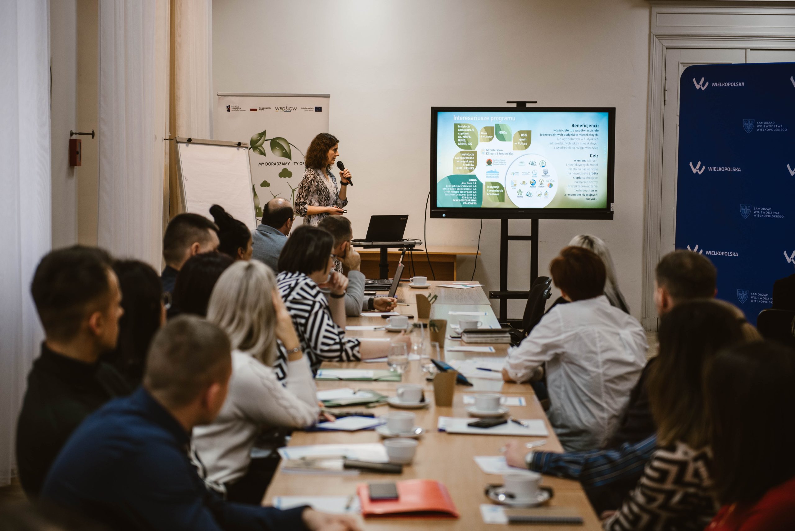 Widok na salkę szkoleniową - grupa ludzi siedzi wzdłuż długiego stołu i słucha prelekcji osoby szkolącej.