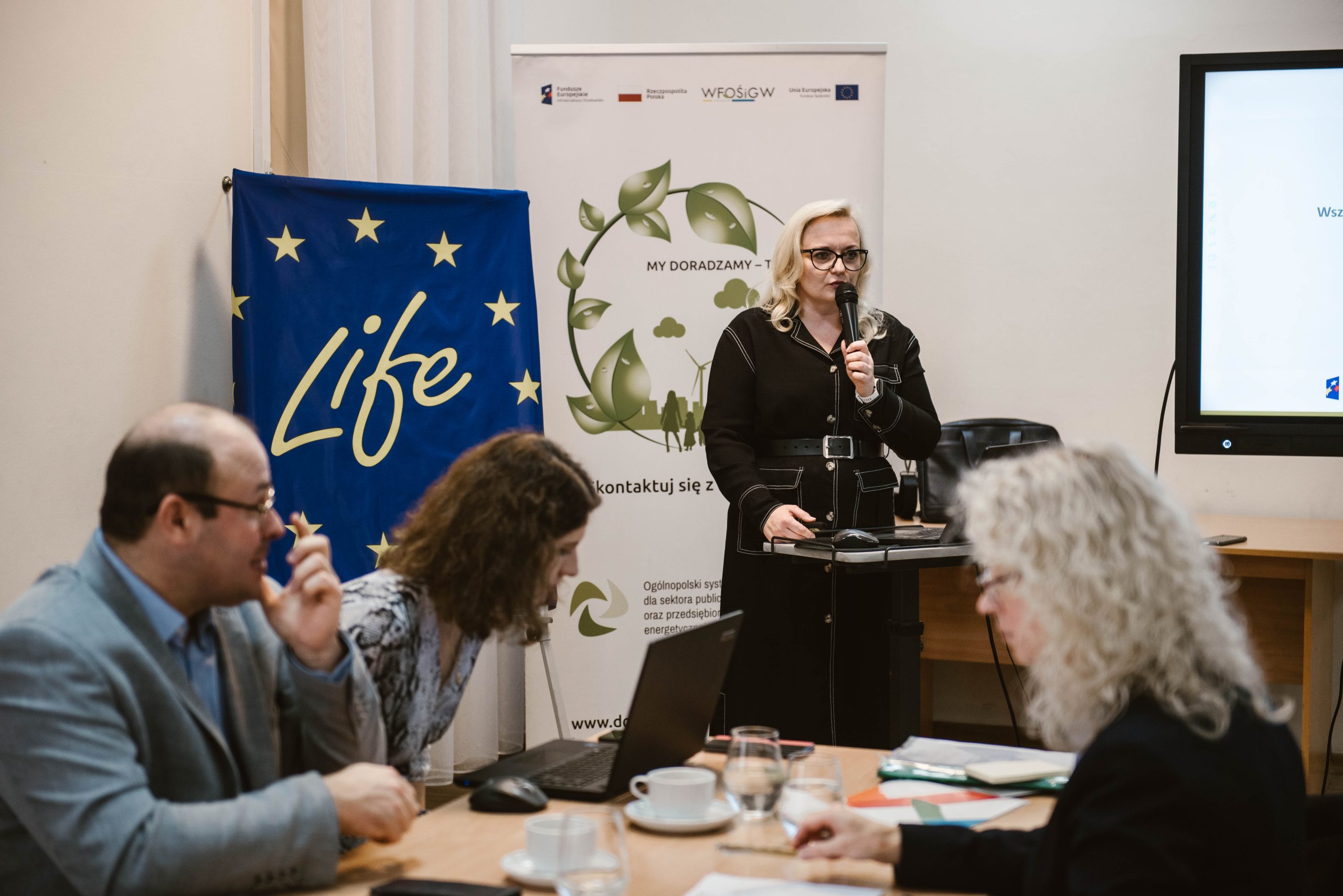 1 osoba prowadzi szkolenie, a uczestnicy siedzą przy długim stole i słuchają i notują.