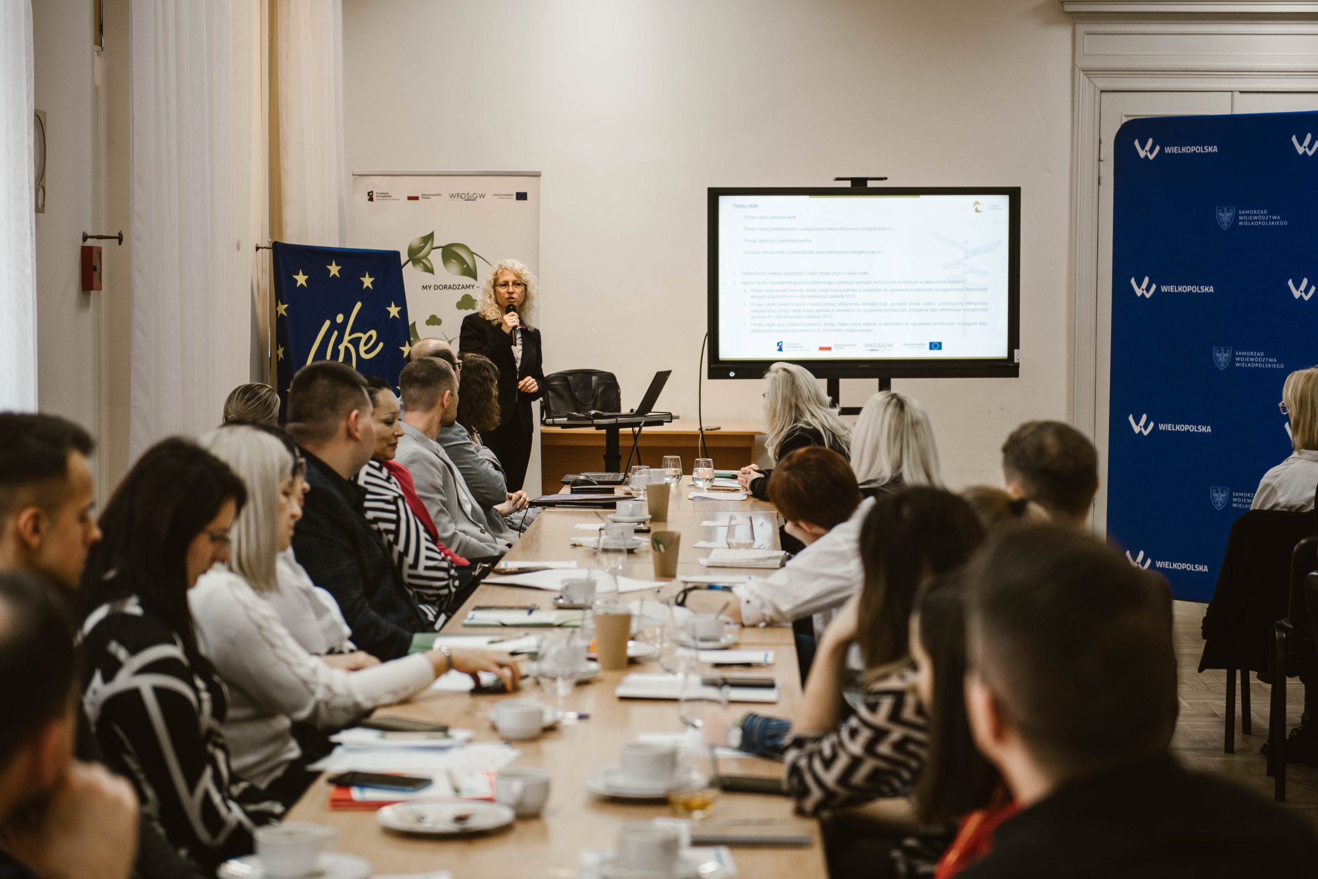 Grupa osób siedzi po obu stronach długiego stołu, jedna osoba prowadzi szkolenie.