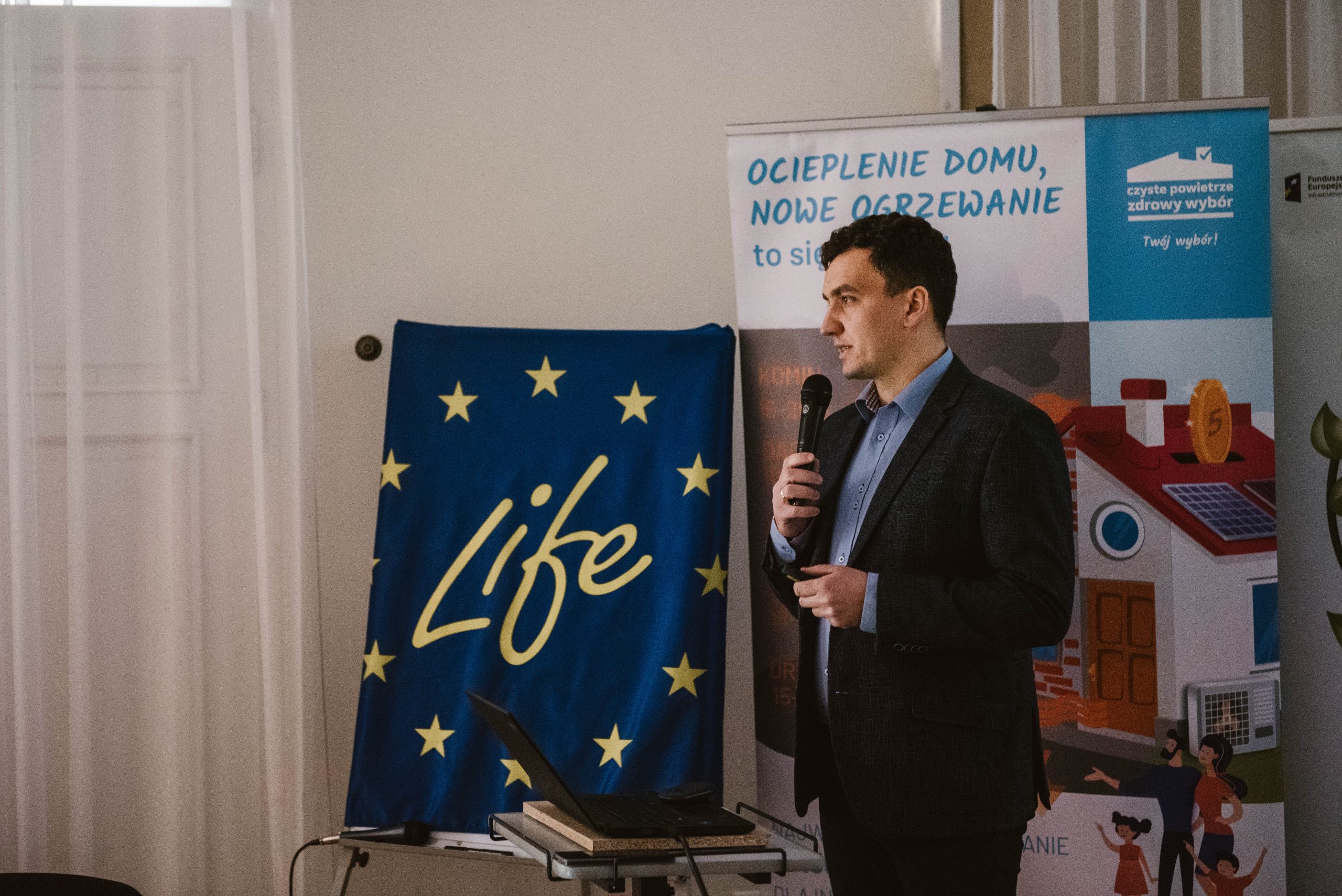 1 osoba stoi na środku sali i przemawia przez mikrofon.
