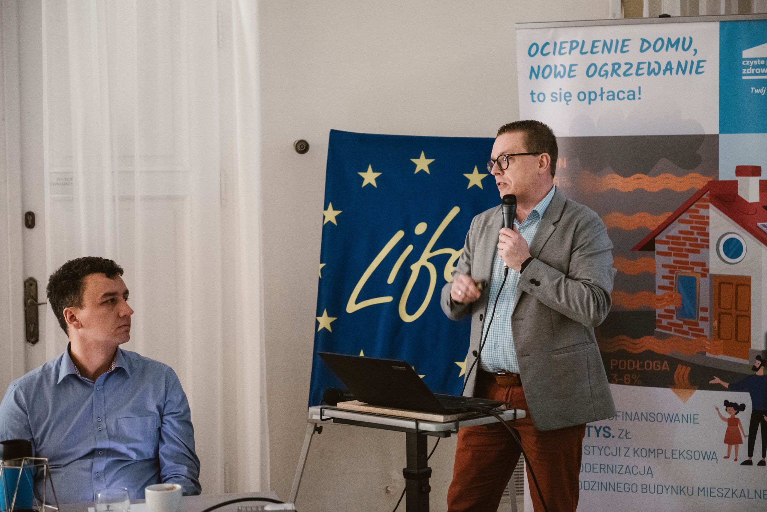 1 osoba stoi na środku sali i przemawia przez mikrofon.