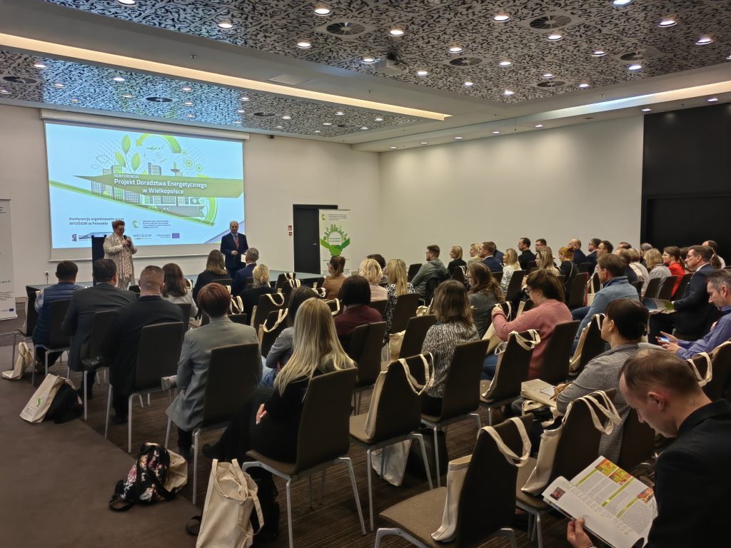 Widok na salę konferencyjną pełną ludzi siedzących na krzesłach, na scenie ekran z prezentacją i jedna osoba prezentuje.