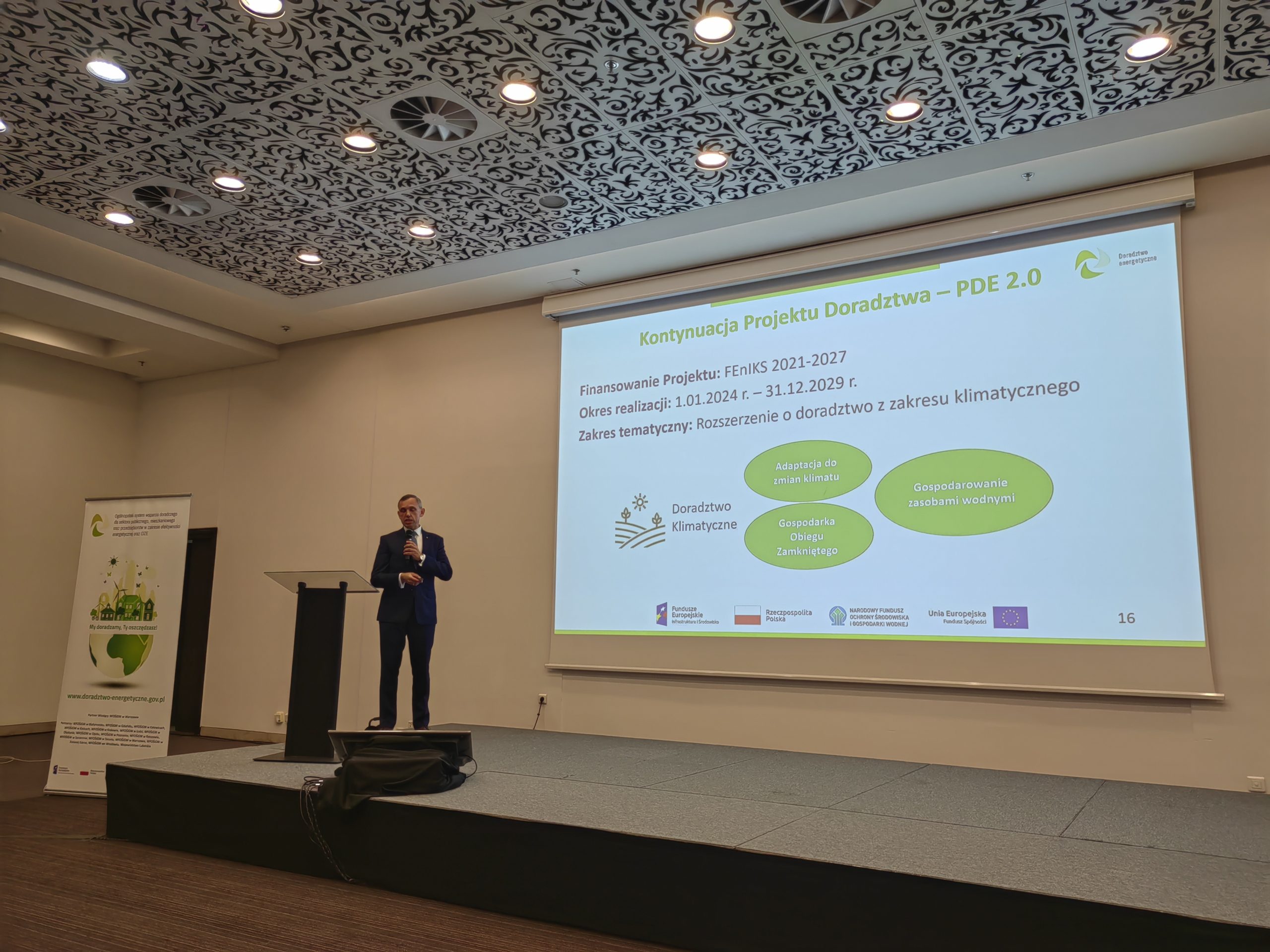 1 osoba stoi na scenie w trakcie konferencji i przemawia do uczestników.