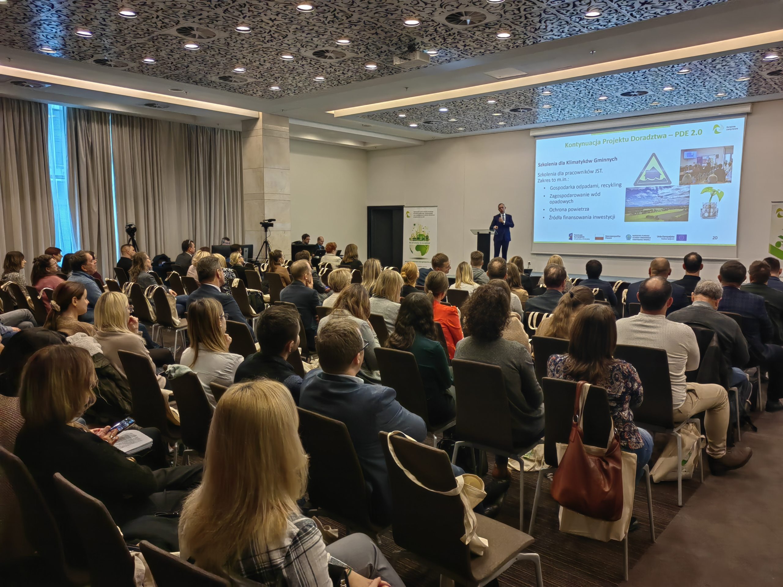 Widok na salę pełną ludzi i jedną osobę przemawiającą ze sceny.
