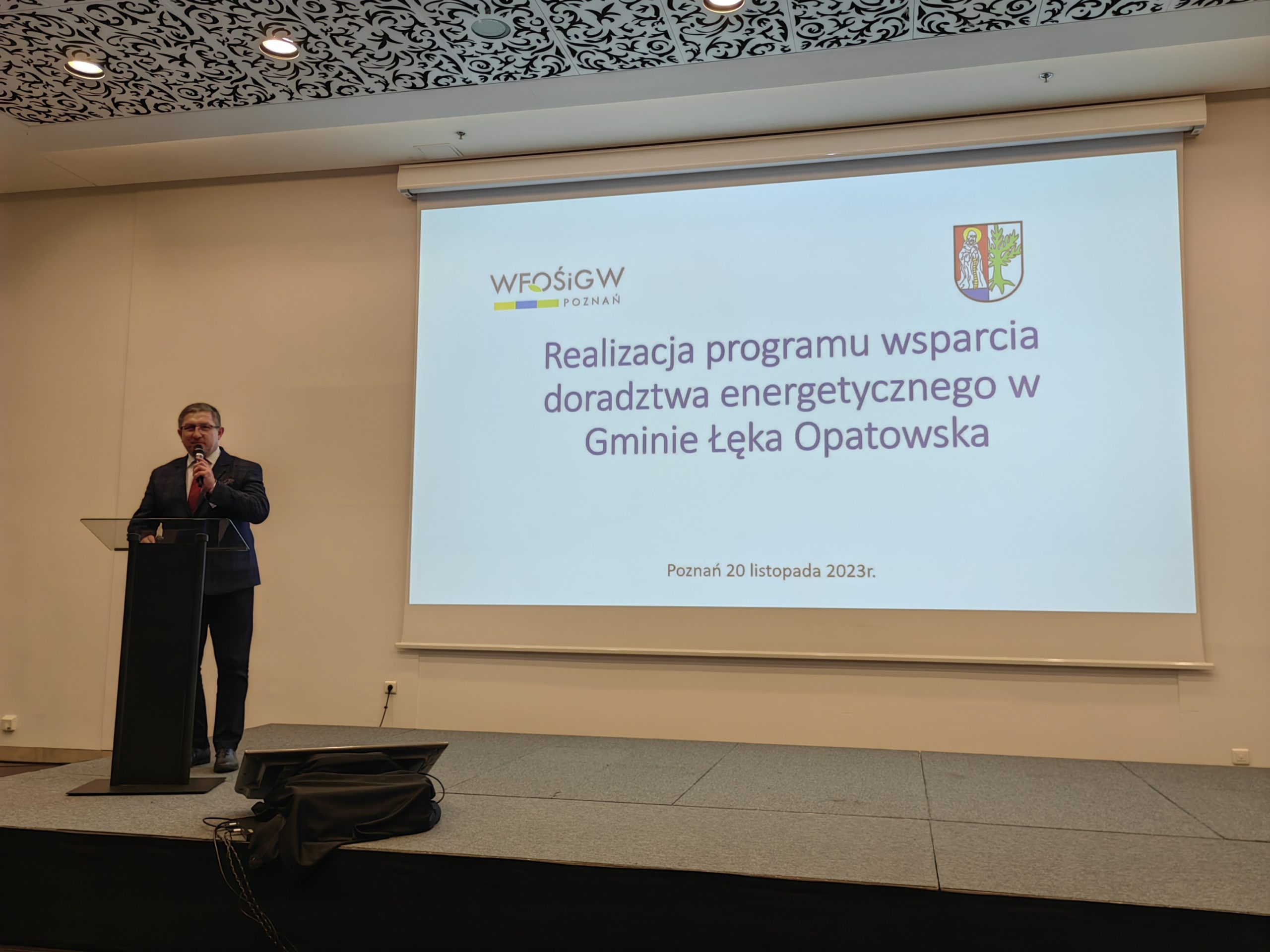1 osoba stoi na scenie w trakcie konferencji i przemawia do uczestników.