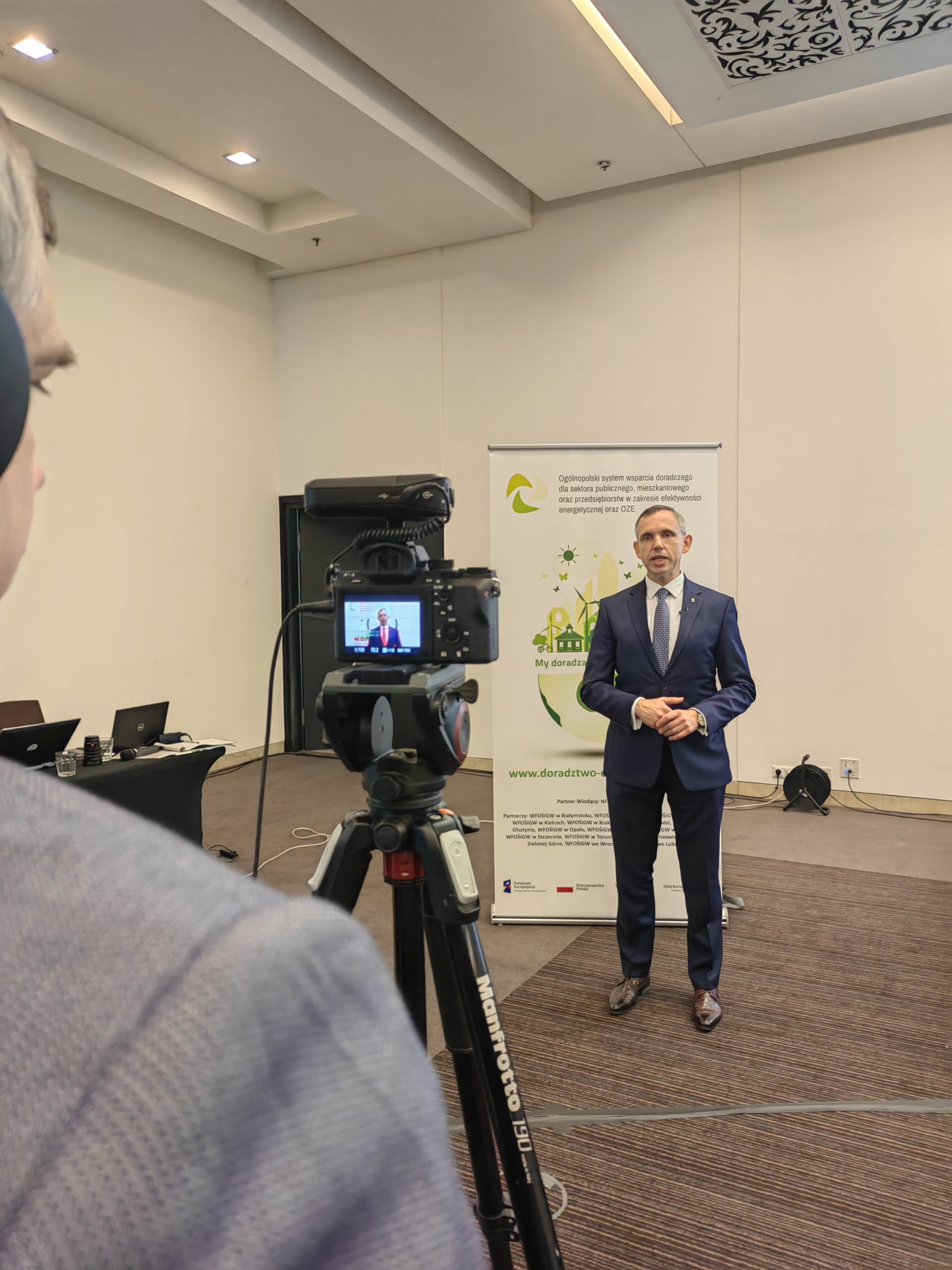 1 osoba stoi na sali konferencji i udziela wywiadu przed kamerą.