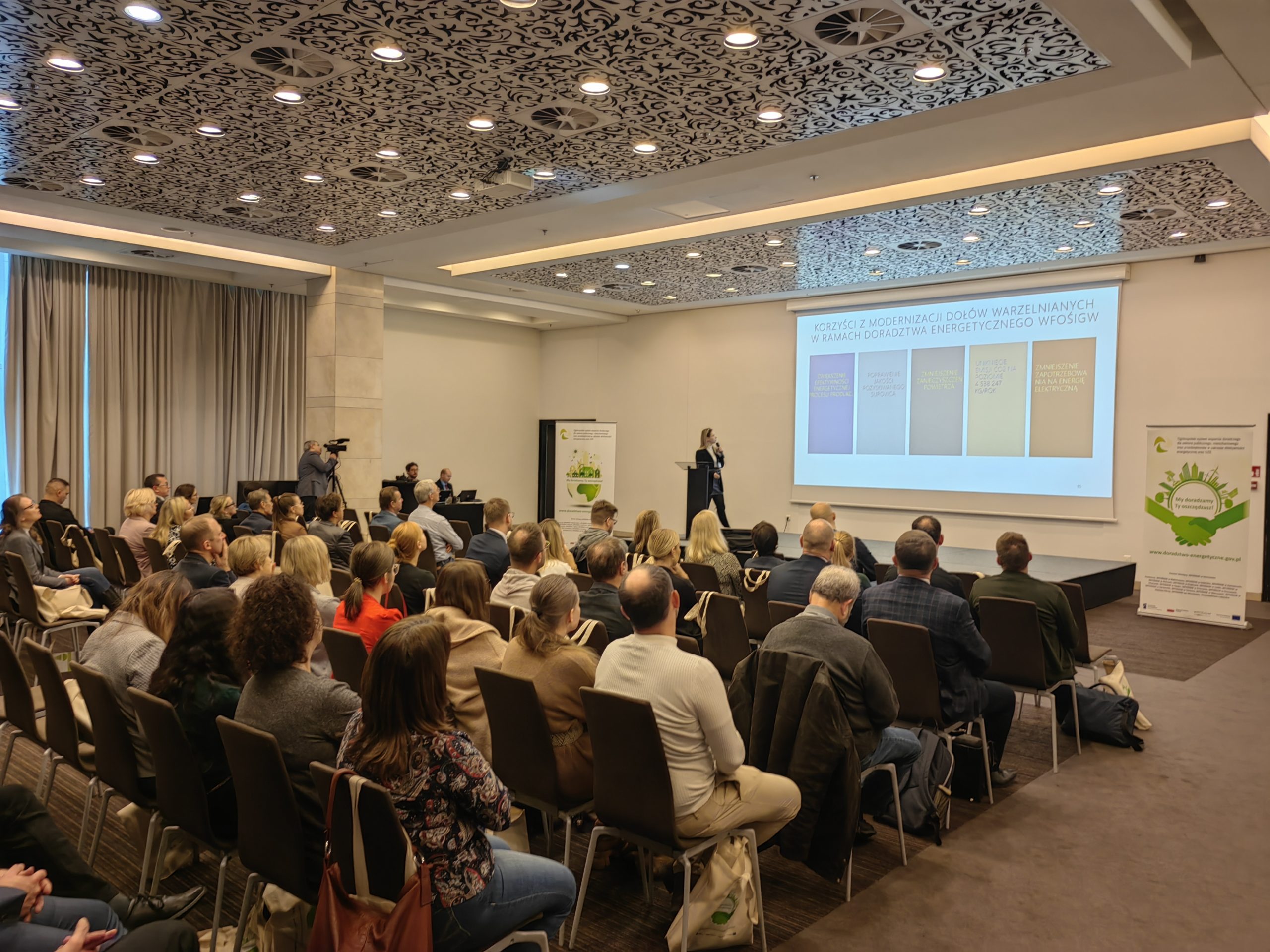 1 osoba stoi na scenie w trakcie konferencji i przemawia do uczestników.