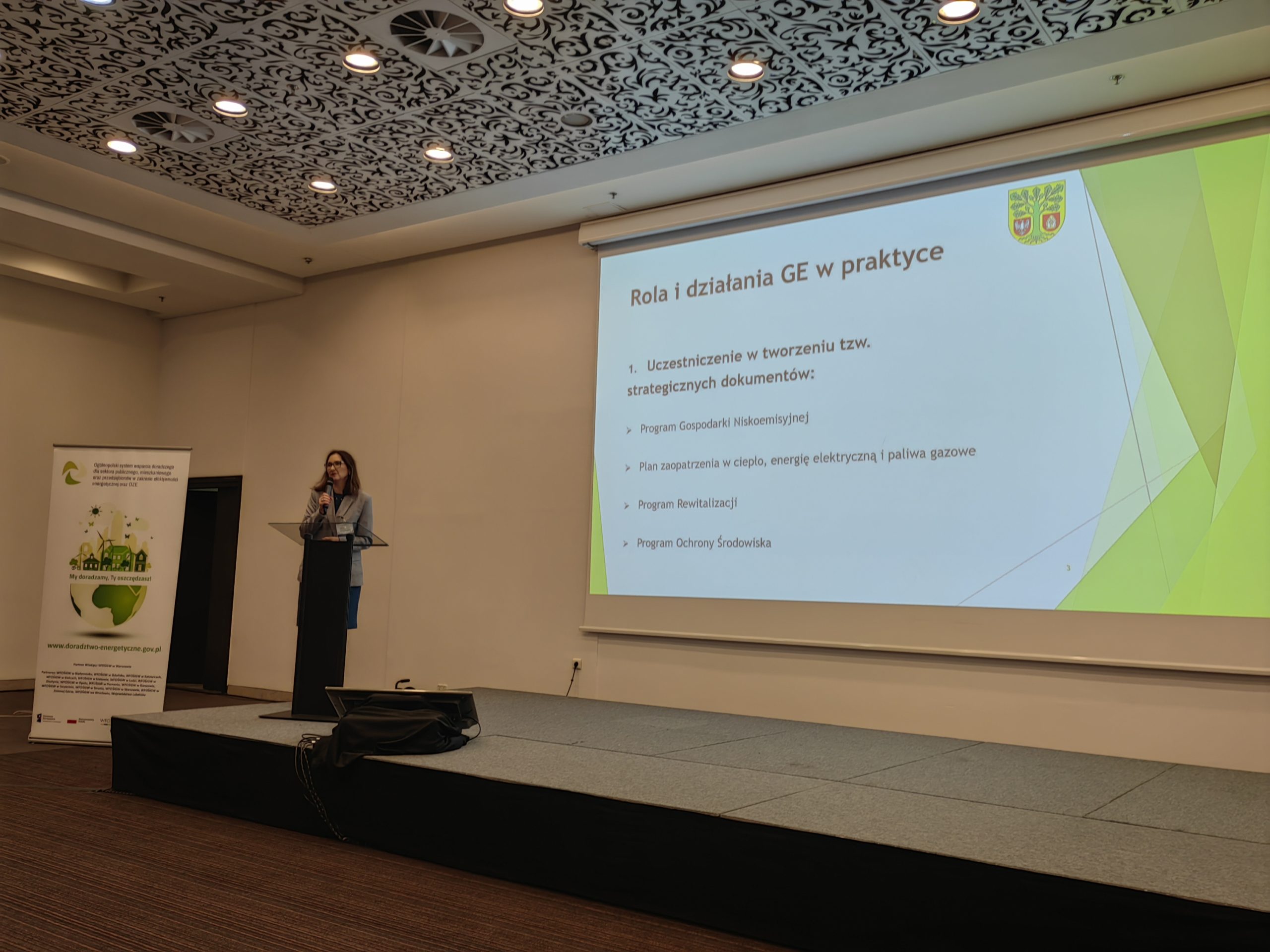 1 osoba stoi na scenie w trakcie konferencji i przemawia do uczestników.